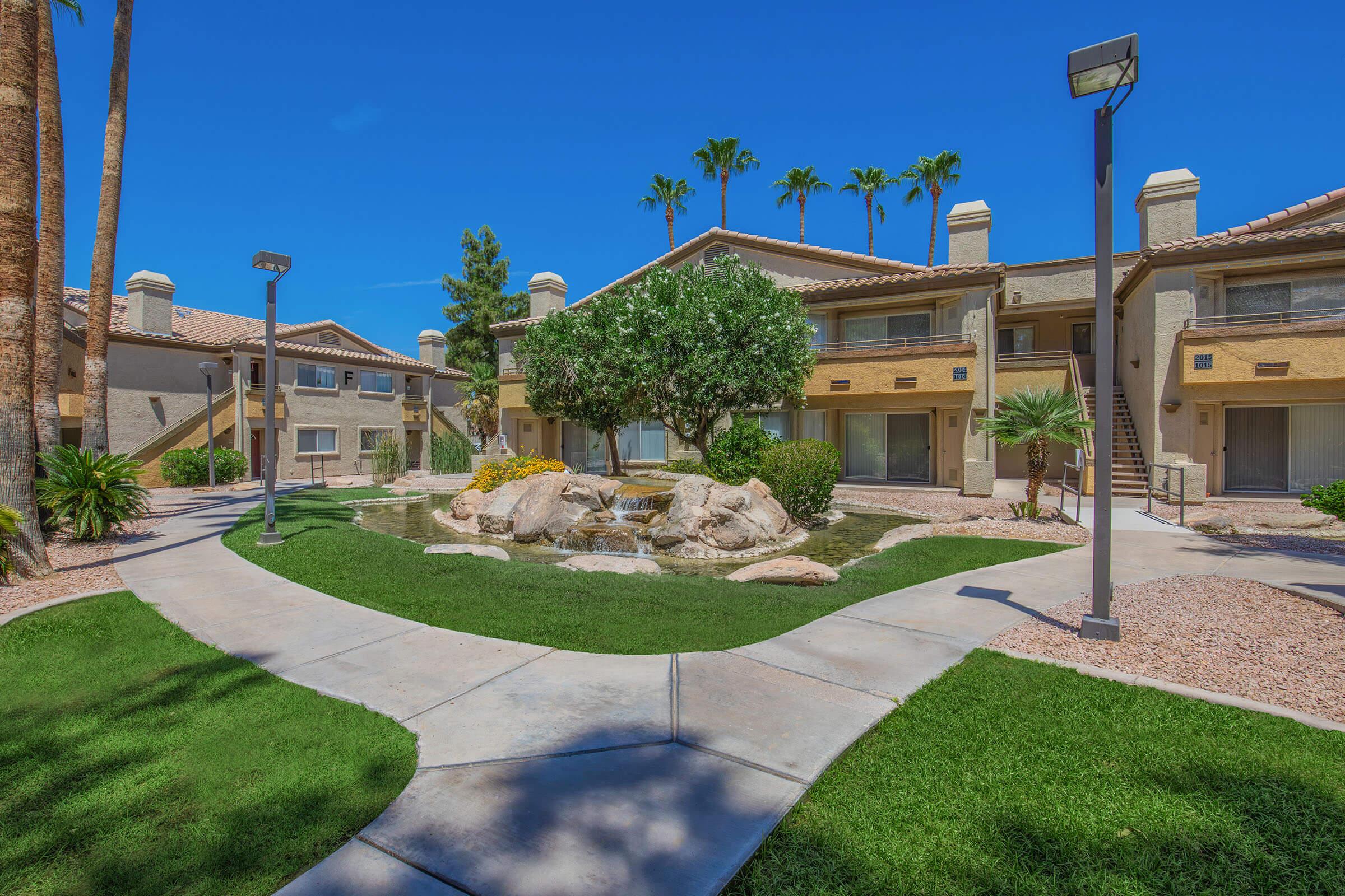 A landscaped courtyard featuring a small rock fountain, surrounded by palm trees and well-maintained grass. Several two-story apartment buildings are visible in the background under a clear blue sky. Pathways connect the buildings, enhancing the community feel.