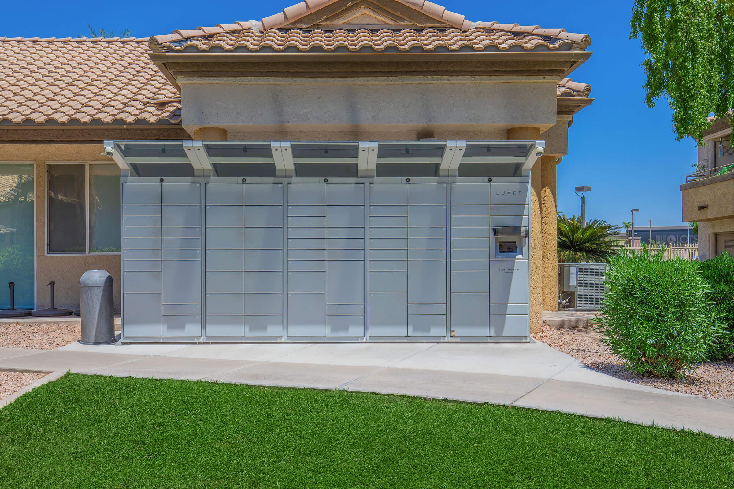 A modern package delivery box installed outside a residential building, featuring multiple compartments for secure storage, with a sleek design and a roof overhead. The structure sits on a landscaped area with green grass and pebbles, accompanied by a view of a palm tree and the building's exterior.