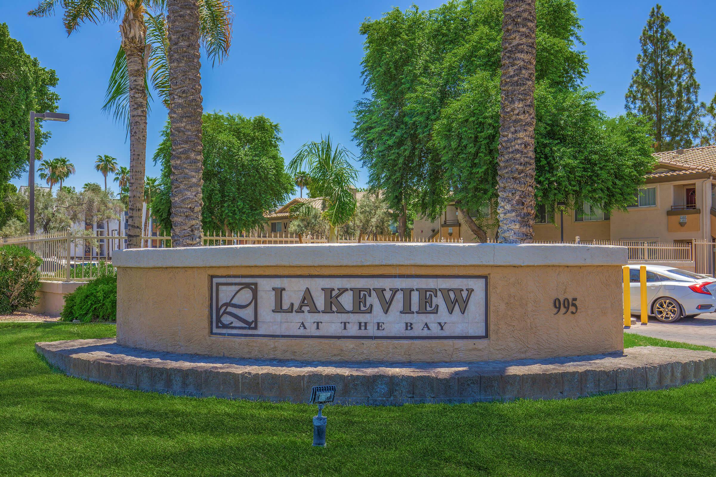 A stone sign displaying "Lakeview at the Bay" with the number "995" beneath it, surrounded by palm trees and lush green grass. The background features residential buildings under a clear blue sky.
