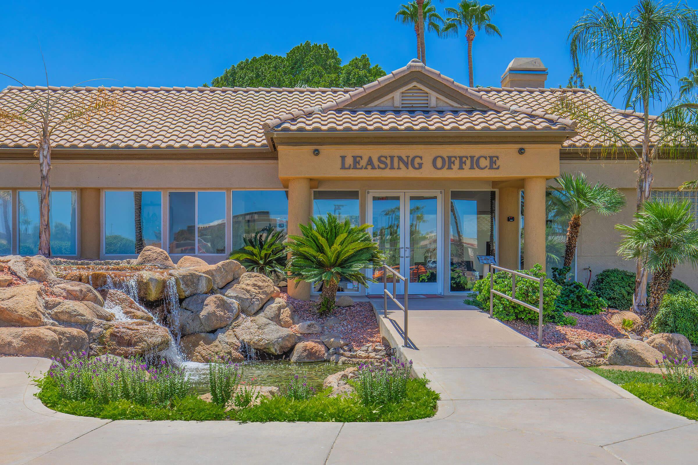 Leasing office building with a tan exterior and a tiled roof. The entrance features large glass doors and tropical plants, beside a rock waterfall and landscaped gardens. Clear blue sky and palm trees are visible in the background, creating a welcoming atmosphere.