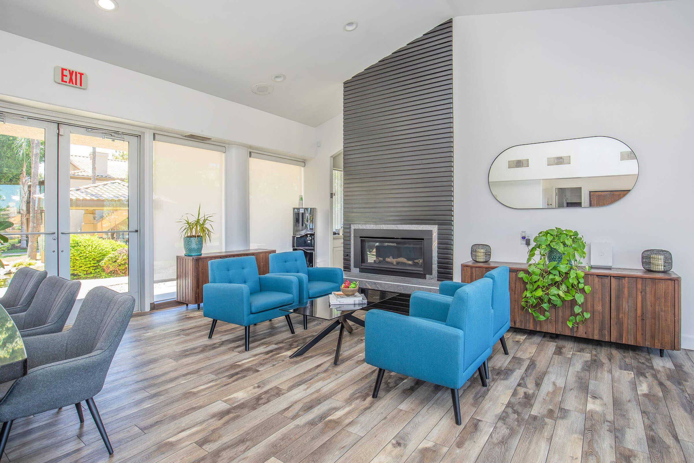 A modern indoor living space featuring two blue armchairs, a black coffee table, and a light wooden floor. There is a stylish mirror on the wall, a plant on a cabinet, and a fireplace. Large windows allow natural light, creating a warm and inviting atmosphere.