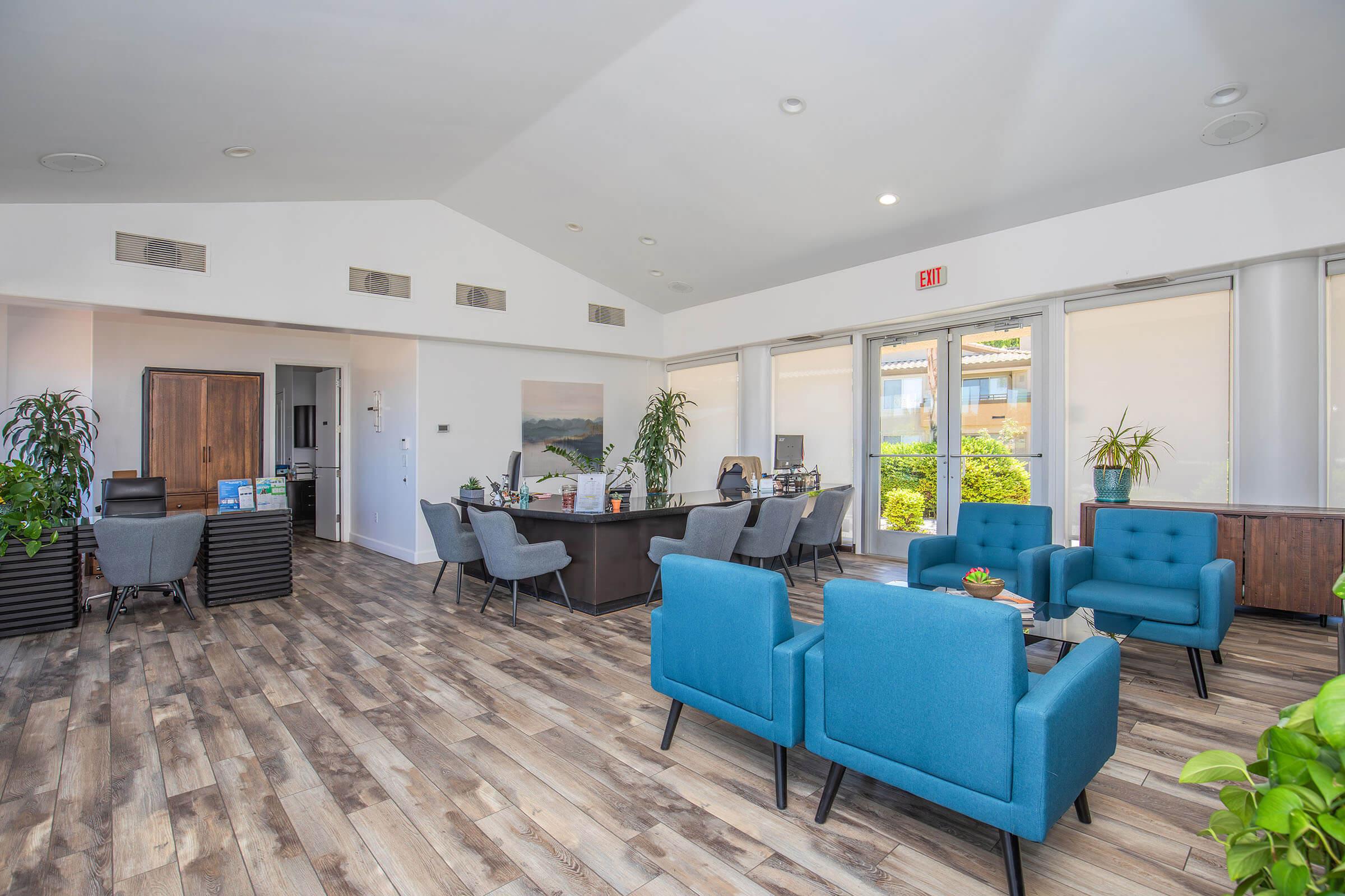 A modern office interior featuring blue chairs and a dark wooden desk. The space has large windows allowing natural light, plants for decoration, and wooden flooring. There are several workstations in the background, creating a professional and inviting atmosphere.