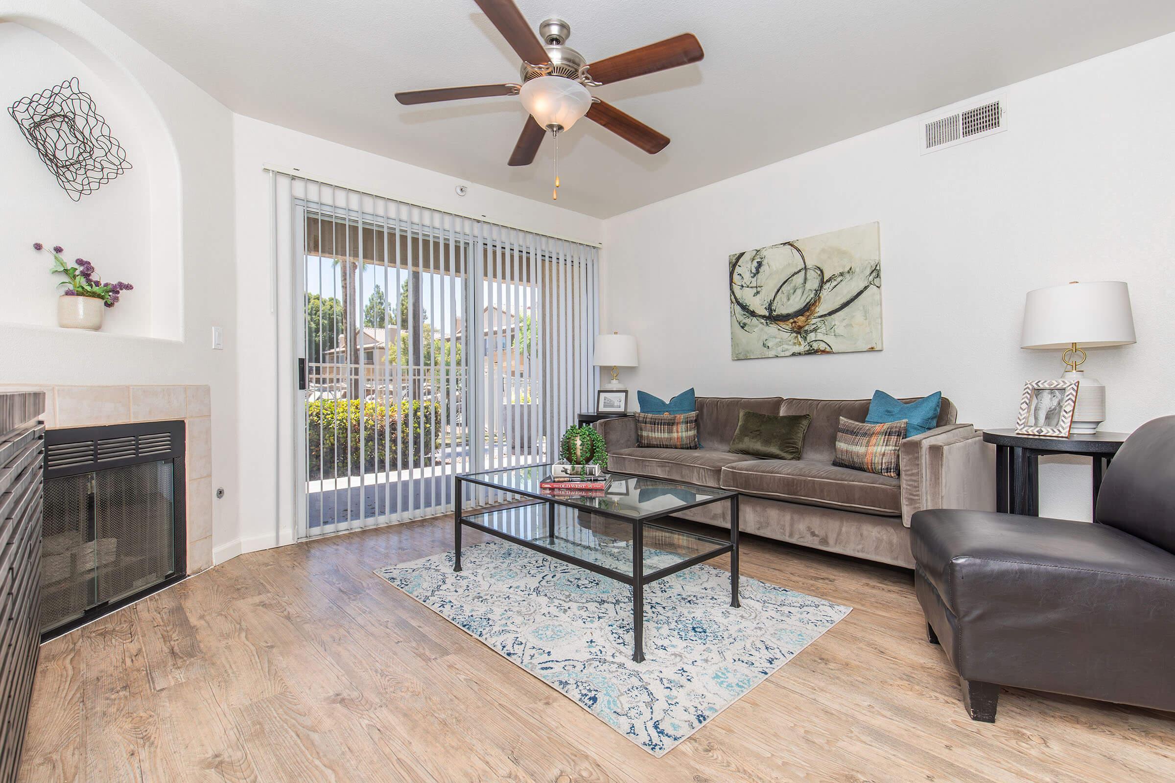 A modern living room featuring a ceiling fan, a comfortable brown sofa with decorative pillows, a glass coffee table on a patterned rug, and two lamps on side tables. A fireplace is to the left, and large sliding glass doors open to a patio with greenery outside.