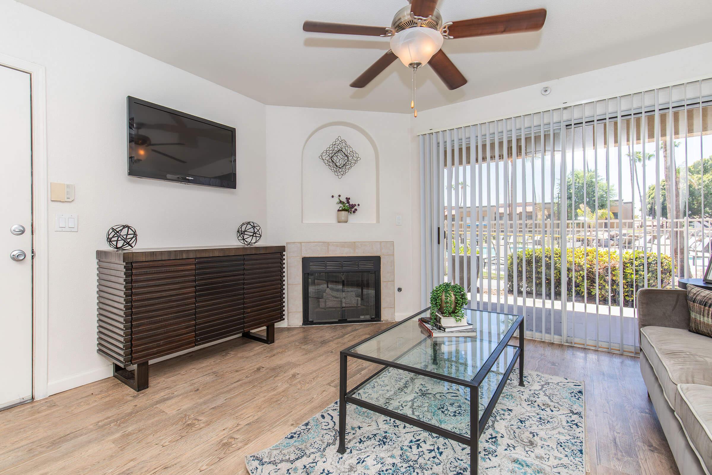 A modern living room featuring a TV mounted on the wall, a stylish wooden console, a cozy fireplace, and large windows with vertical blinds letting in natural light. A coffee table sits on a decorative rug, complementing the comfortable sofa and overall contemporary decor.
