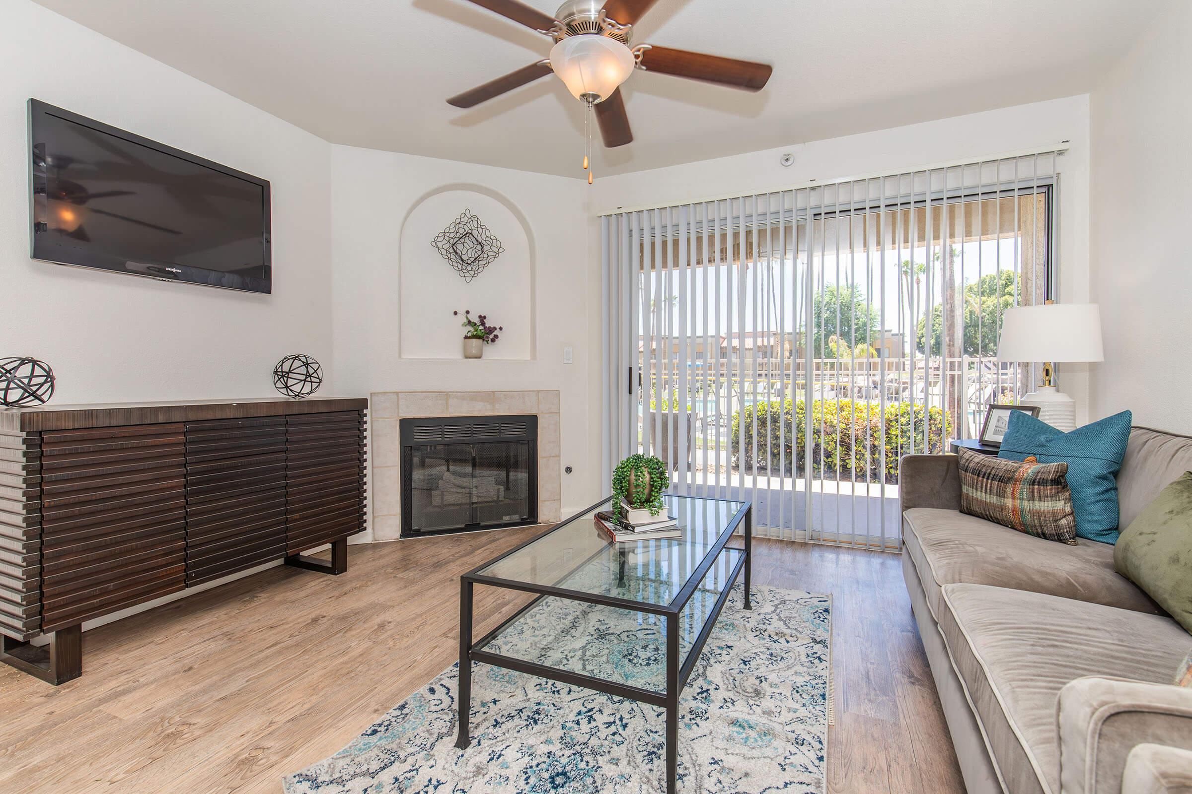A cozy living room with a flat-screen TV mounted on the wall, a stylish wooden media console, a glass coffee table on a patterned rug, and a comfortable sofa adorned with decorative pillows. There are sliding glass doors leading to an outdoor area, allowing natural light to fill the space.