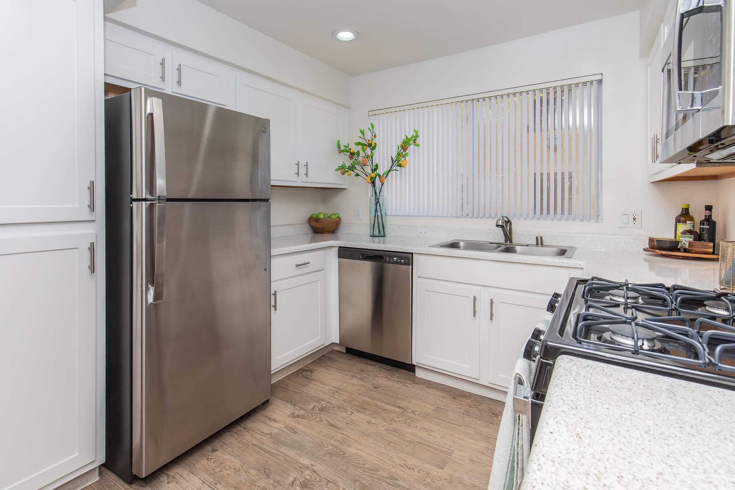 Modern kitchen featuring stainless steel appliances, including a refrigerator and dishwasher. The kitchen showcases white cabinets, a sink under a window with blinds, and a countertop. A vase with flowers is displayed on the counter, enhancing the bright and clean environment.