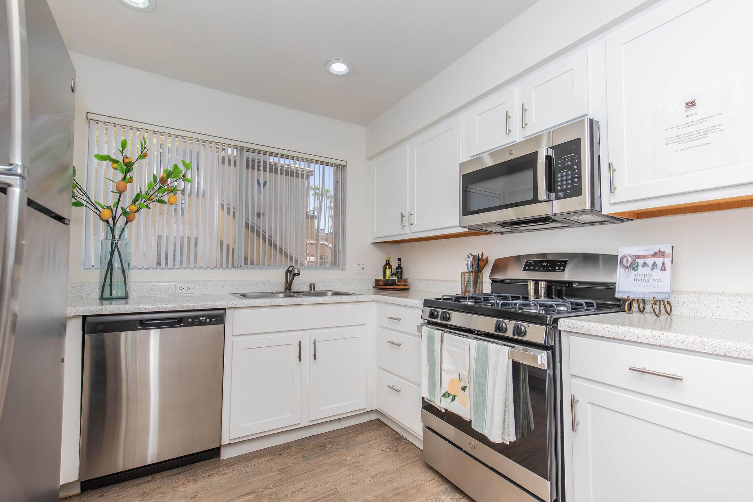 Modern kitchen featuring stainless steel appliances, including a dishwasher and oven. There's a window with blinds, bright white cabinetry, and a countertop with a light speckled design. Decorative vase with flowers sits by the window, creating a welcoming atmosphere.
