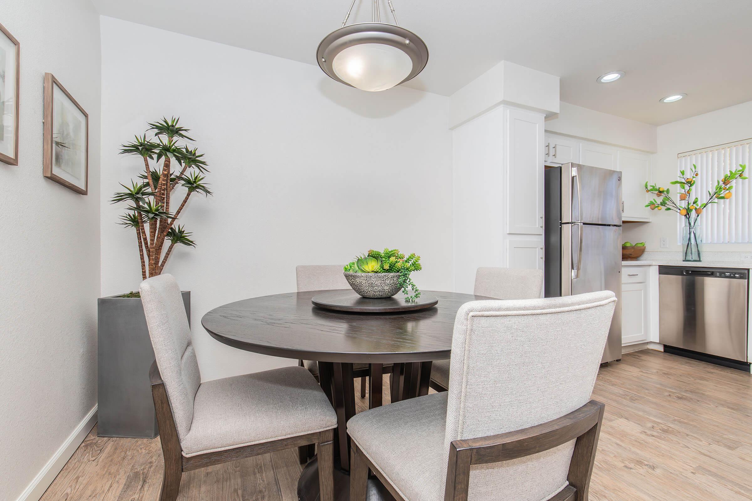 A modern dining area featuring a round wooden table surrounded by four light-colored upholstered chairs. A decorative bowl filled with greenery sits at the center of the table. In the background, there are potted plants and kitchen appliances visible, contributing to a bright and fresh ambiance.