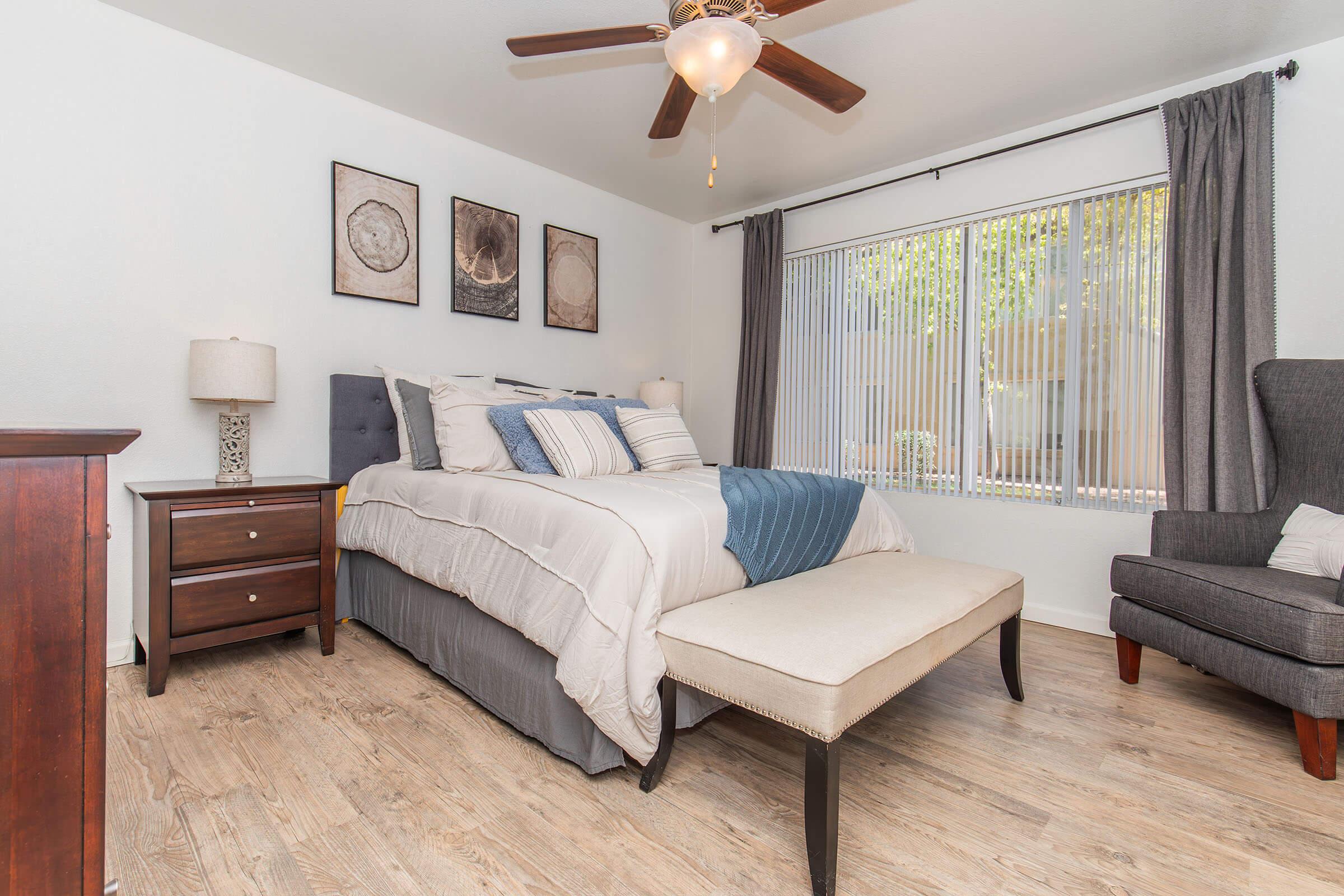 A cozy bedroom featuring a queen-sized bed with soft linens and decorative pillows. There's a stylish bench at the foot of the bed, a wooden nightstand with a lamp, and a comfortable armchair. Natural light streams in through a large window, accompanied by beige curtains, while framed artwork adorns the wall.