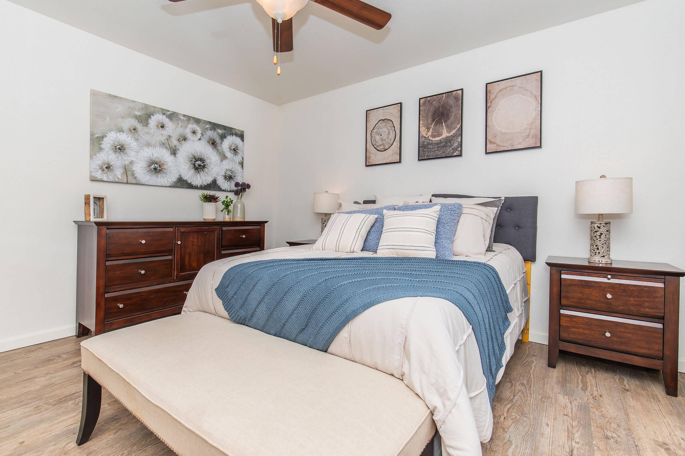 A cozy bedroom featuring a neatly made bed with a blue throw blanket, flanked by two bedside tables with lamps. A wooden dresser sits opposite the bed, and a piece of wall art depicting flowers hangs above it, complemented by three framed images. The room has a light, airy feel with wooden flooring.