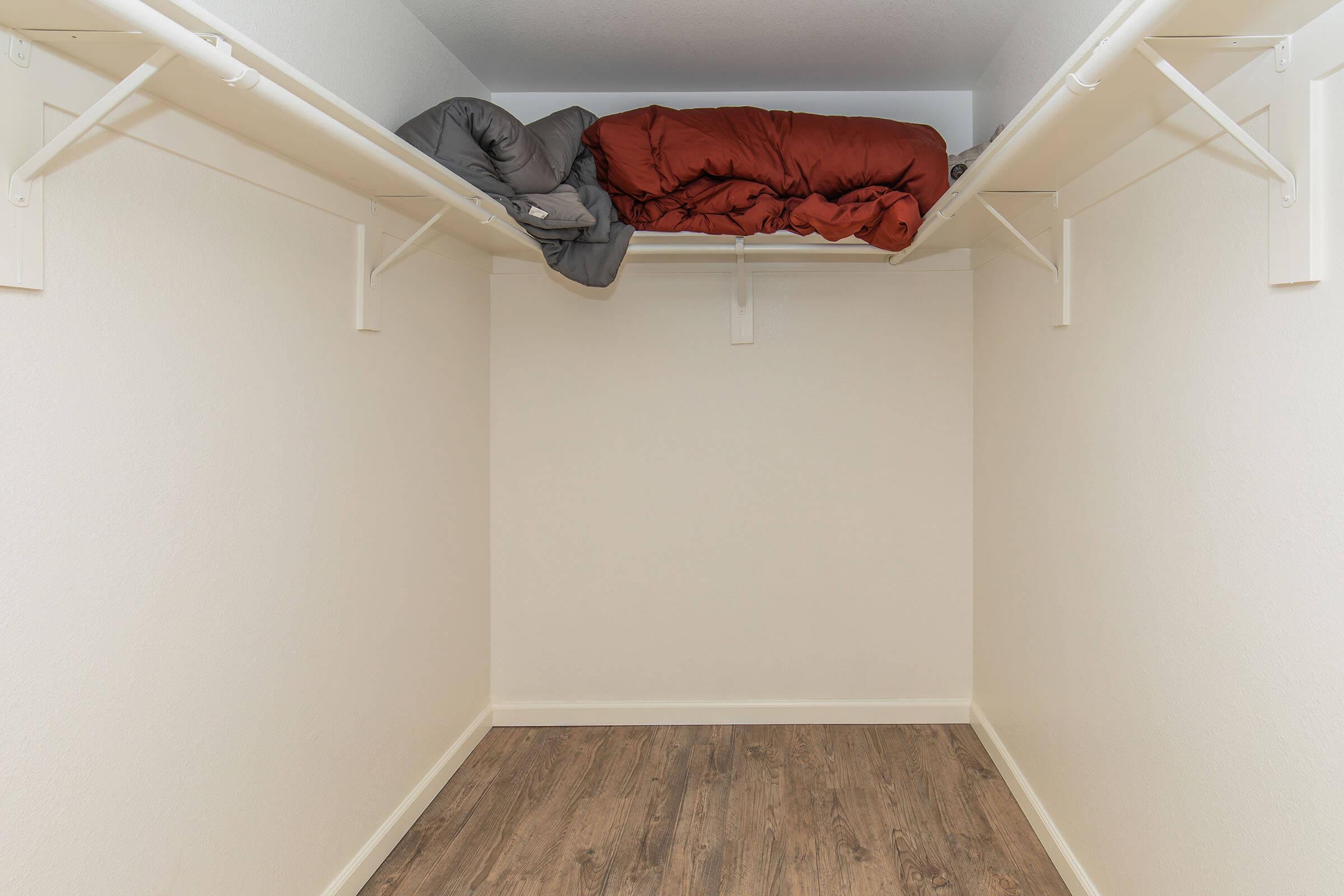 A spacious closet featuring a plain white interior and light-colored wooden flooring. Two shelves are installed, with a red blanket and a grey sleeping bag stacked on the upper shelf. The closet is empty below, providing ample storage space.