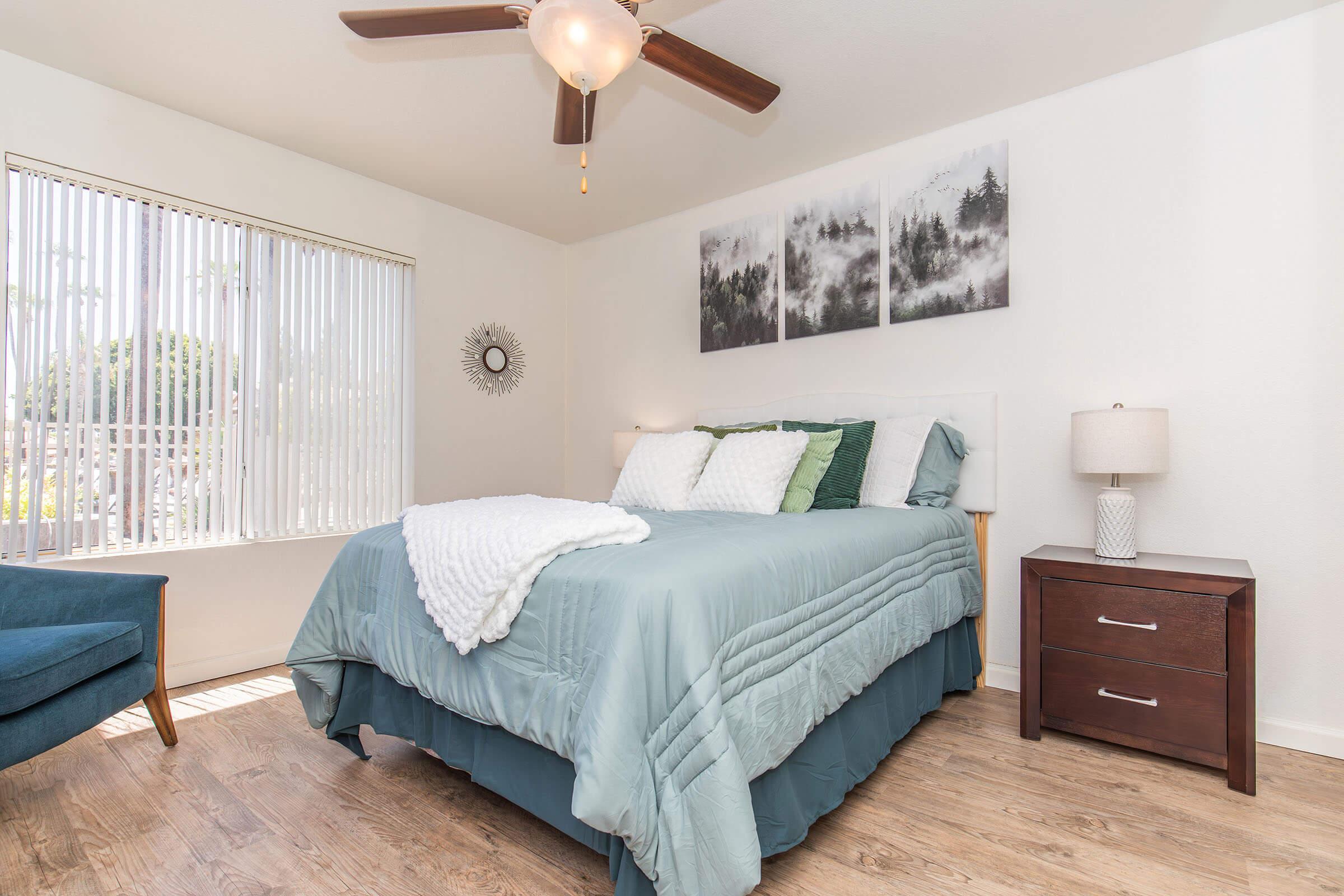 A cozy bedroom featuring a queen-sized bed with light blue bedding, accent pillows, and a white throw. There is a nightstand with a lamp beside the bed, a chair in the corner, and three black and white landscape prints on the wall. Natural light streams in through a window with vertical blinds.