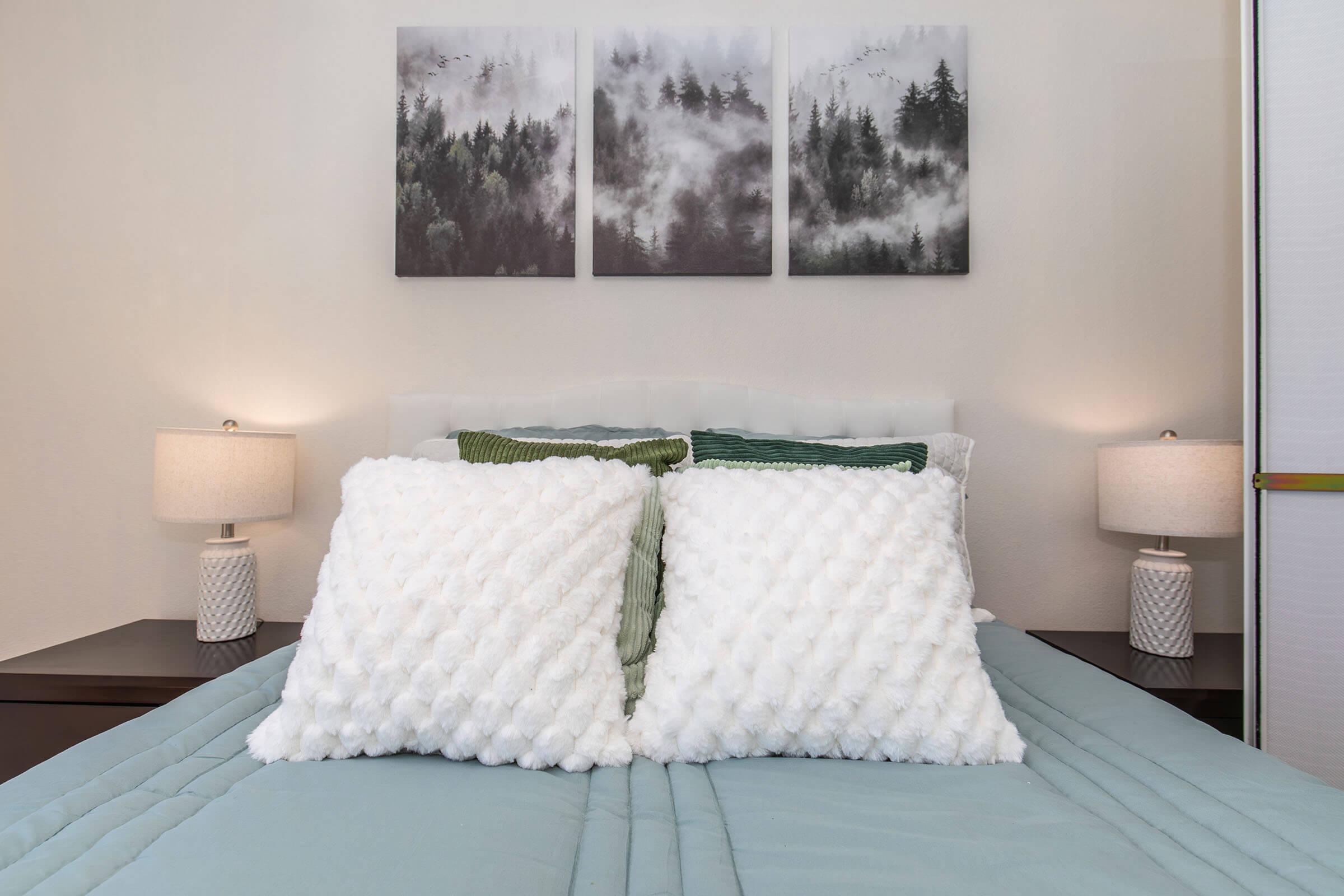 A neatly made bed with a light blue duvet, featuring two large fluffy white pillows and a green accent pillow. On either side of the bed, there are modern lamps with textured bases. Above the bed, three framed landscape prints show misty mountains and forests, adding a serene touch to the room's decor.