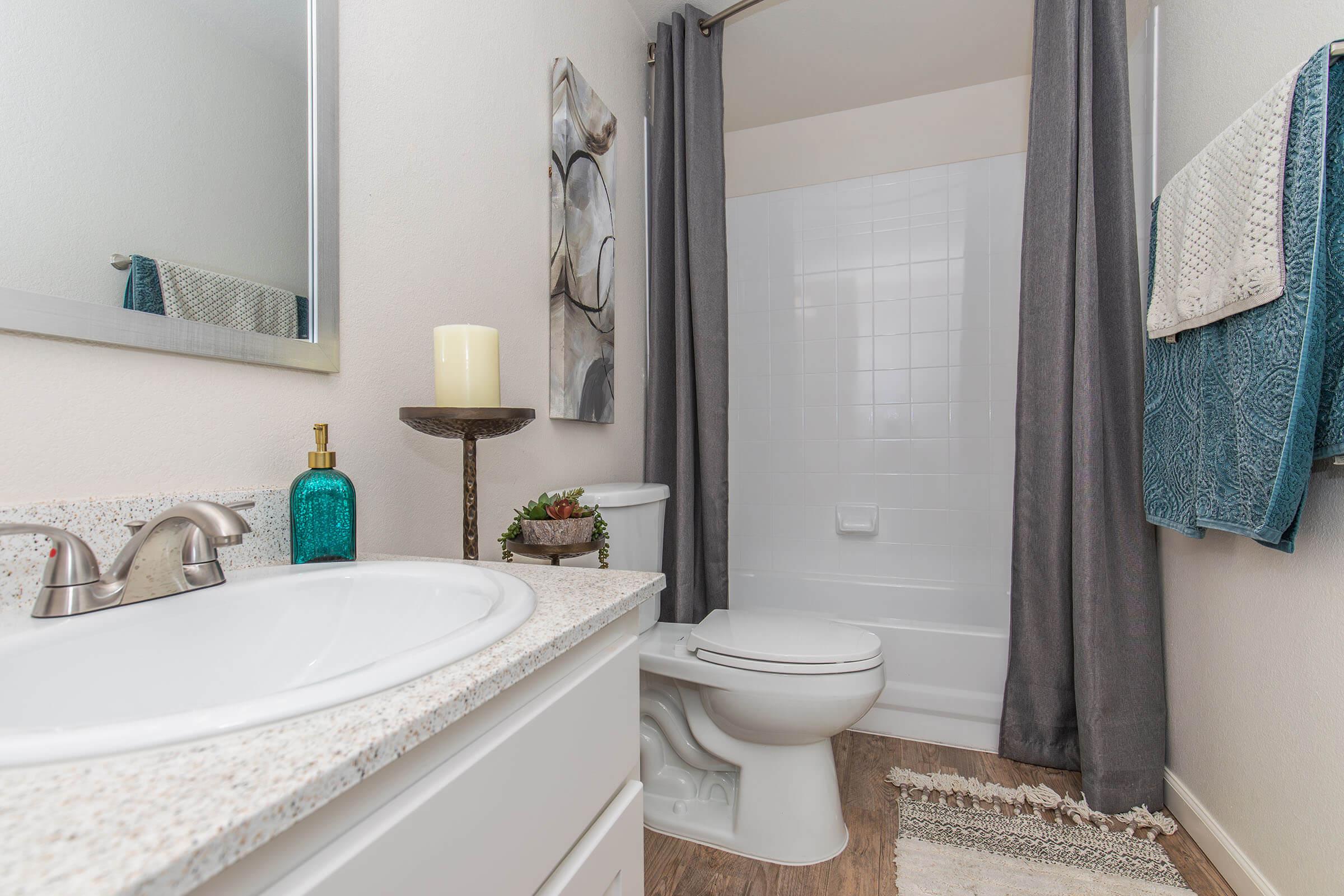 A clean and modern bathroom featuring a white bathtub with a shower, gray curtains, a white sink with a granite countertop, and decorative items like a candle and a teal glass bottle. The walls are light-colored, and there's a framed artwork on display. A woven rug adds texture to the wooden floor.