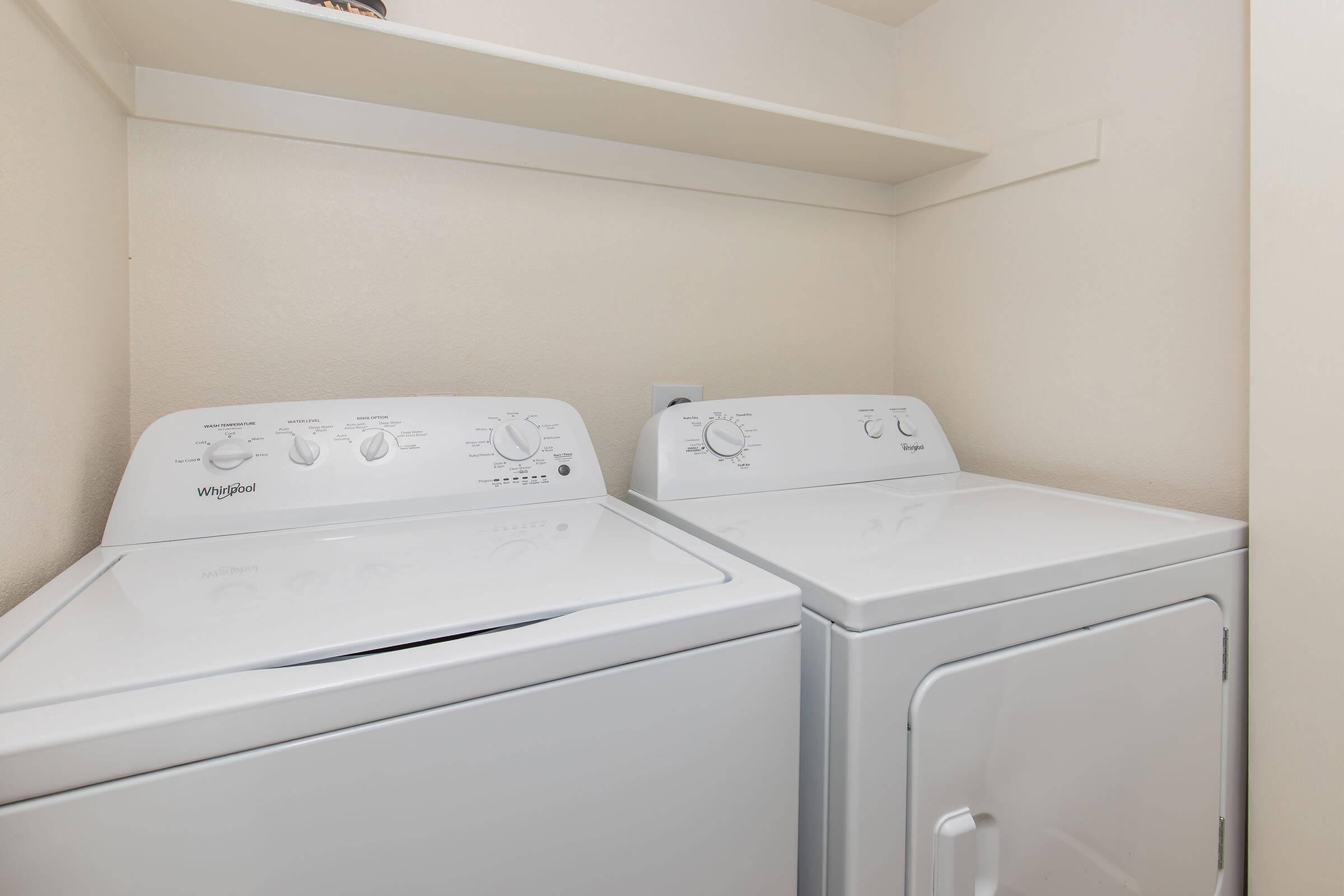 A white washing machine and dryer side by side in a small laundry space, with a shelf above for storage. The machines are clean and well-maintained, set against light-colored walls.