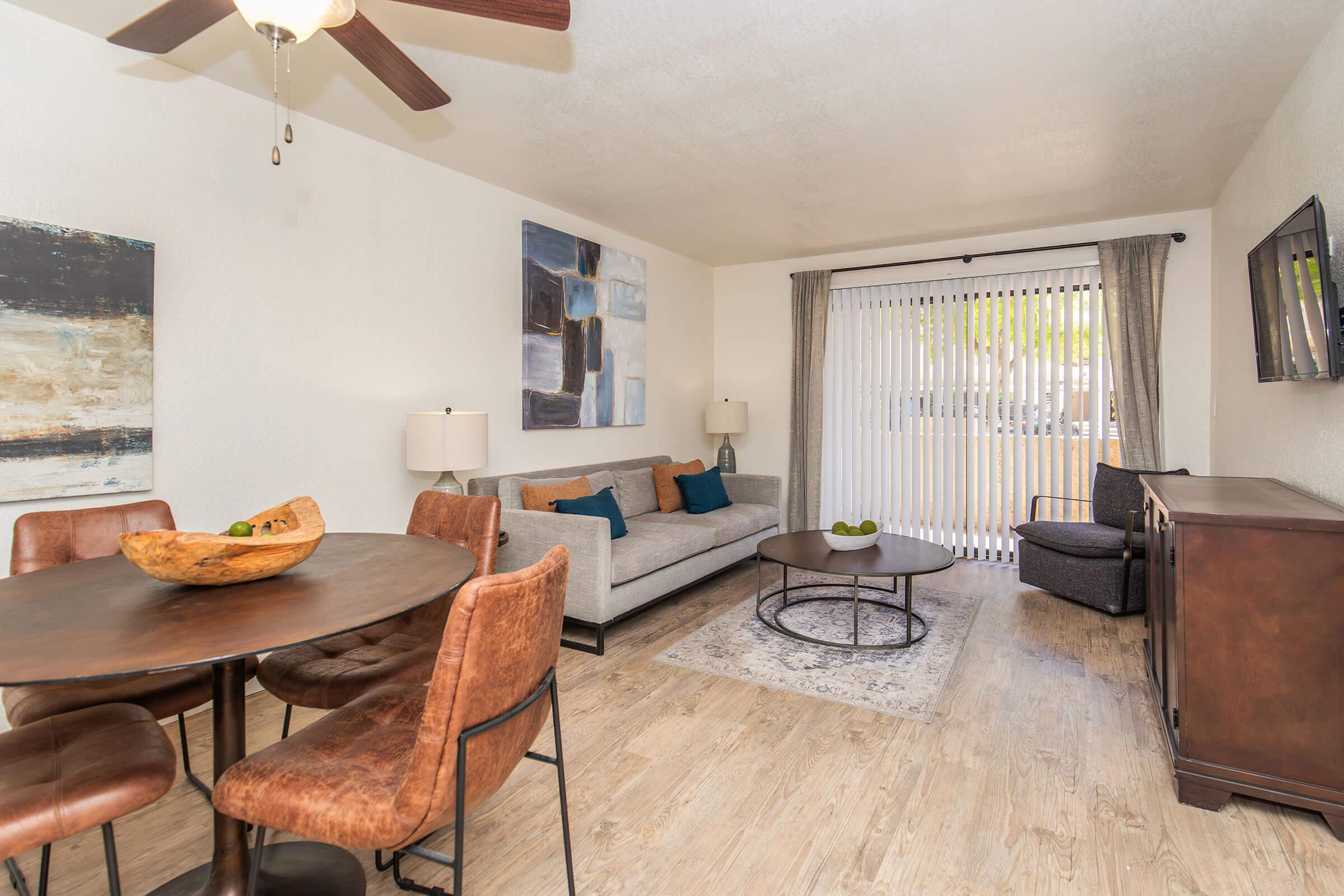 Cozy living space featuring a round dining table with leather chairs, a comfortable gray sofa with colorful cushions, and a modern coffee table. Light streams through large sliding glass doors with vertical blinds. Wall art and a TV add a stylish touch to the room, creating a welcoming atmosphere.