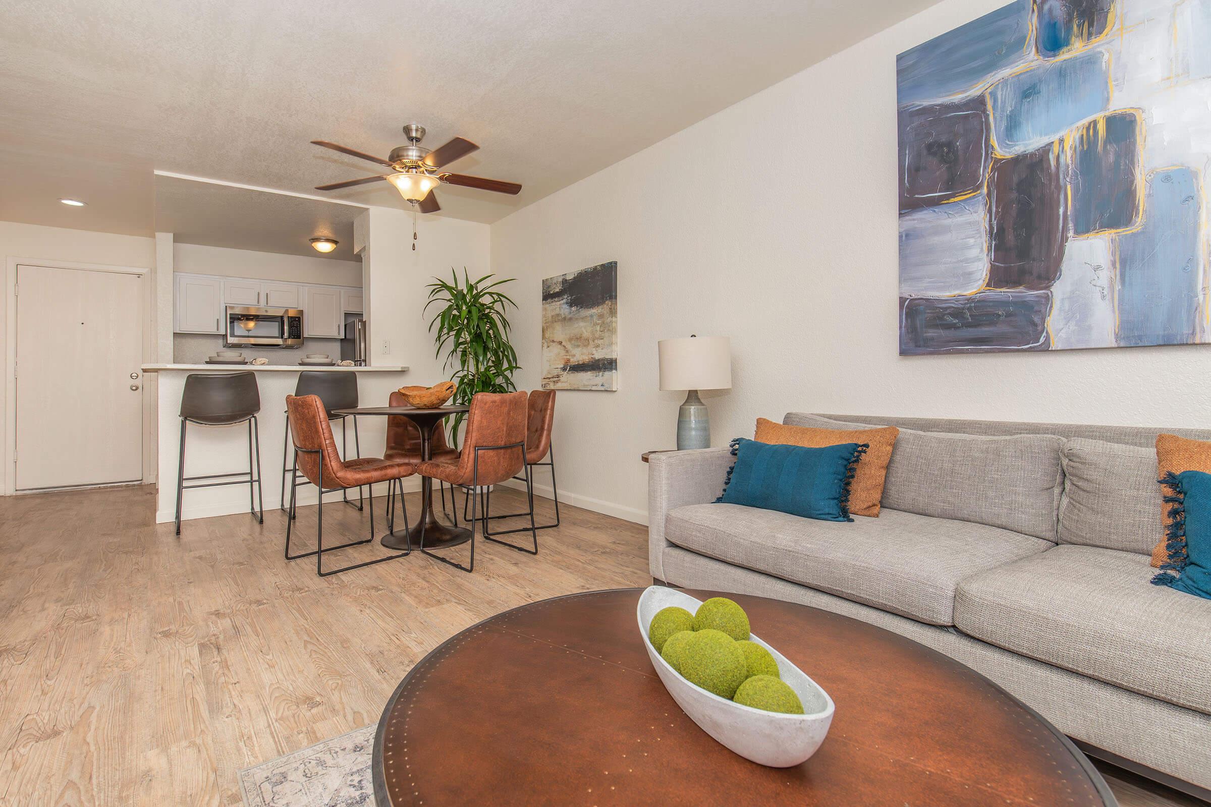 A modern living room featuring a gray sofa with colorful cushions, a round coffee table with green decorative balls, a dining area with metal chairs, and a kitchen in the background. Natural lighting fills the space, highlighted by a ceiling fan and artwork on the walls.