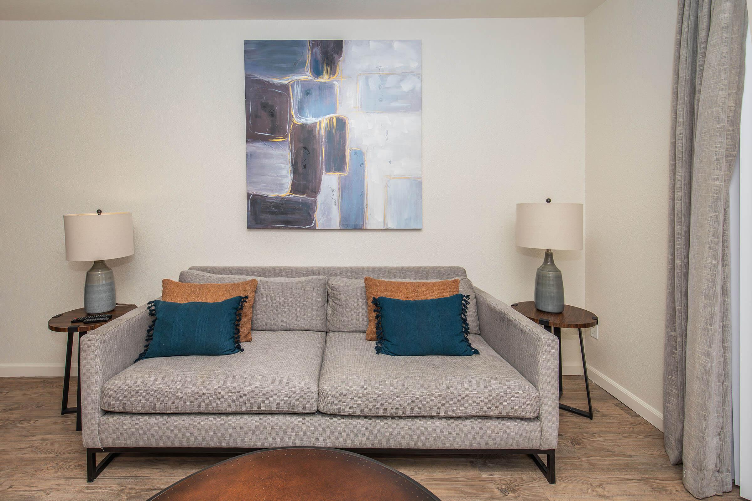 Modern living room with a light gray sofa featuring two blue cushions and two decorative brown pillows. A large abstract painting in shades of blue and white hangs on the white wall above the sofa. There are two side tables with stylish lamps, and a round wooden coffee table in front of the sofa on a wooden floor.
