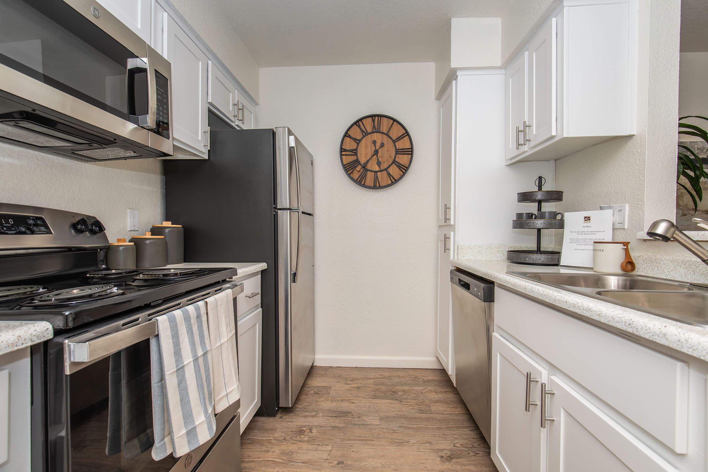 Modern kitchen featuring stainless steel appliances including a range, refrigerator, and dishwasher. White cabinetry and a light countertop create a bright space. A decorative wall clock adds a touch of style, while a plant sits on the counter. Hardwood-like flooring enhances the contemporary look.