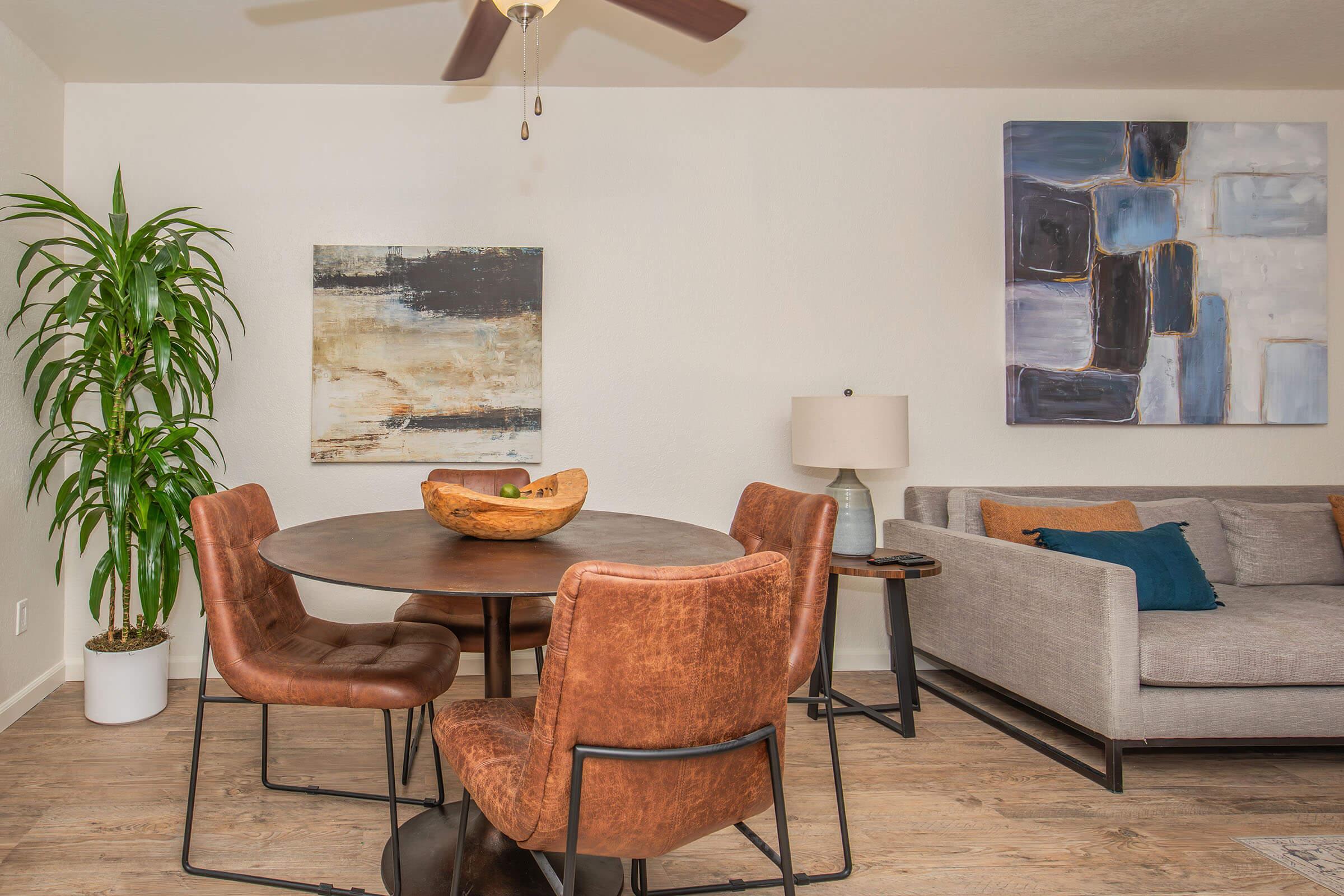 A cozy living space featuring a round dining table with brown chairs, a modern gray sofa with blue accent pillows, a decorative bowl on the table, a tall green plant, and contemporary wall art in muted colors. The room has light wood flooring and a ceiling fan.