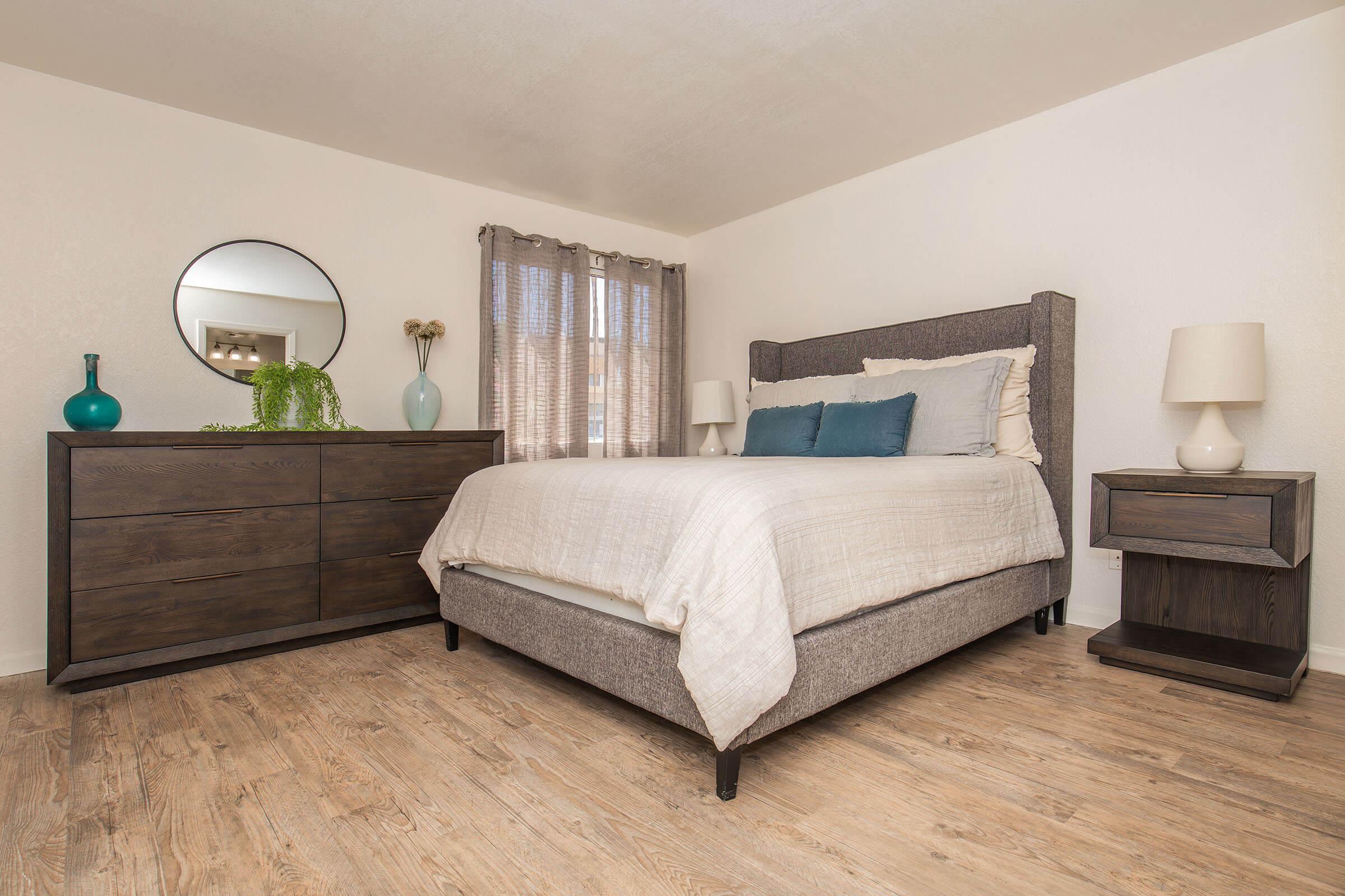 A cozy bedroom featuring a neatly made bed with light bedding and decorative pillows. There's a round mirror on the wall, a wooden dresser with a vase and plant, and a nightstand with a lamp. Soft natural light filters through sheer curtains, highlighting the warm wood flooring.