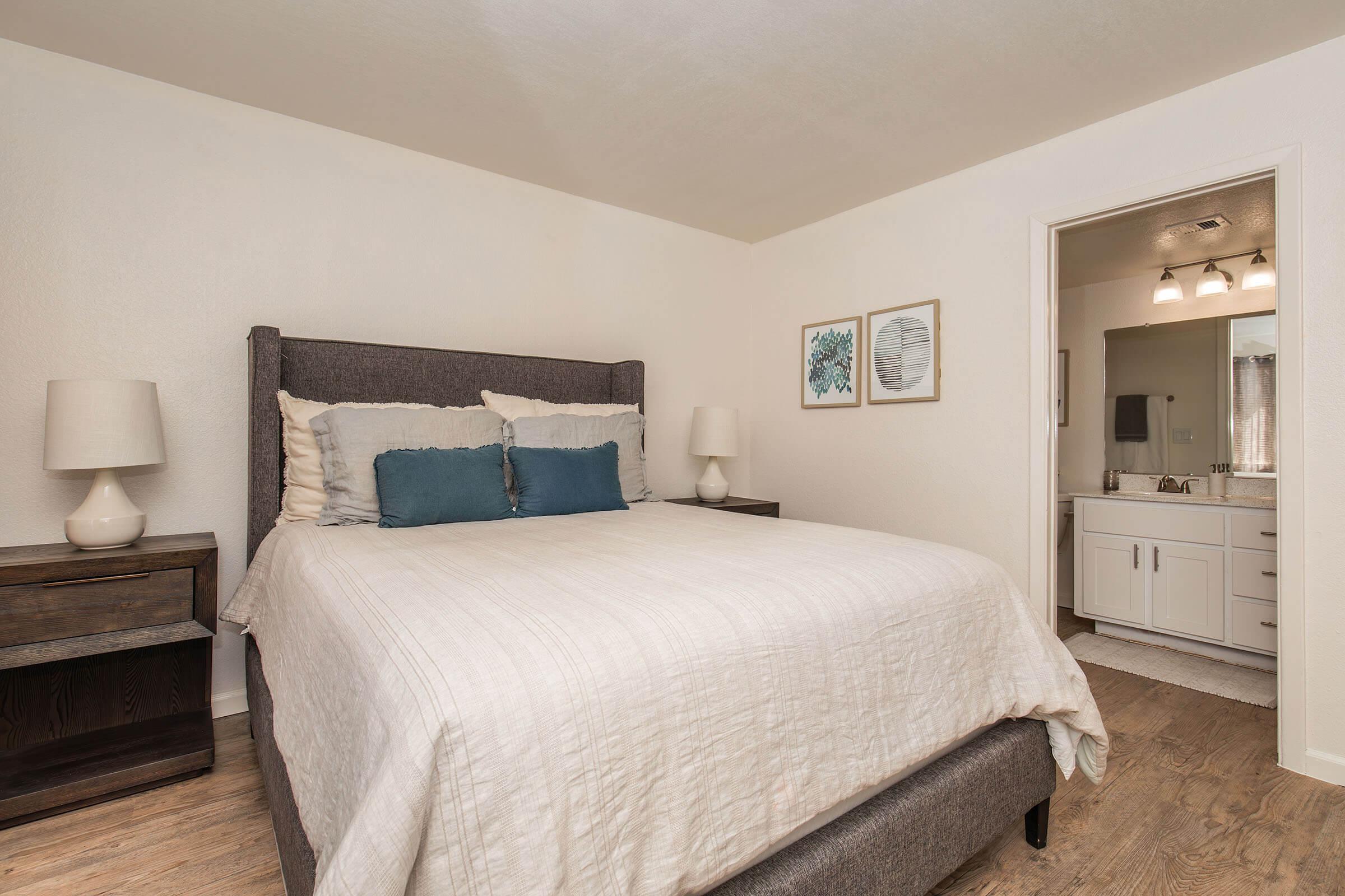 A well-lit and minimalist bedroom featuring a large, neatly made bed with white and blue bedding. There are two bedside lamps on either side and framed artwork on the wall. A doorway leads to a bathroom area with visible lighting fixtures. The flooring is light wood, enhancing the spacious feel.