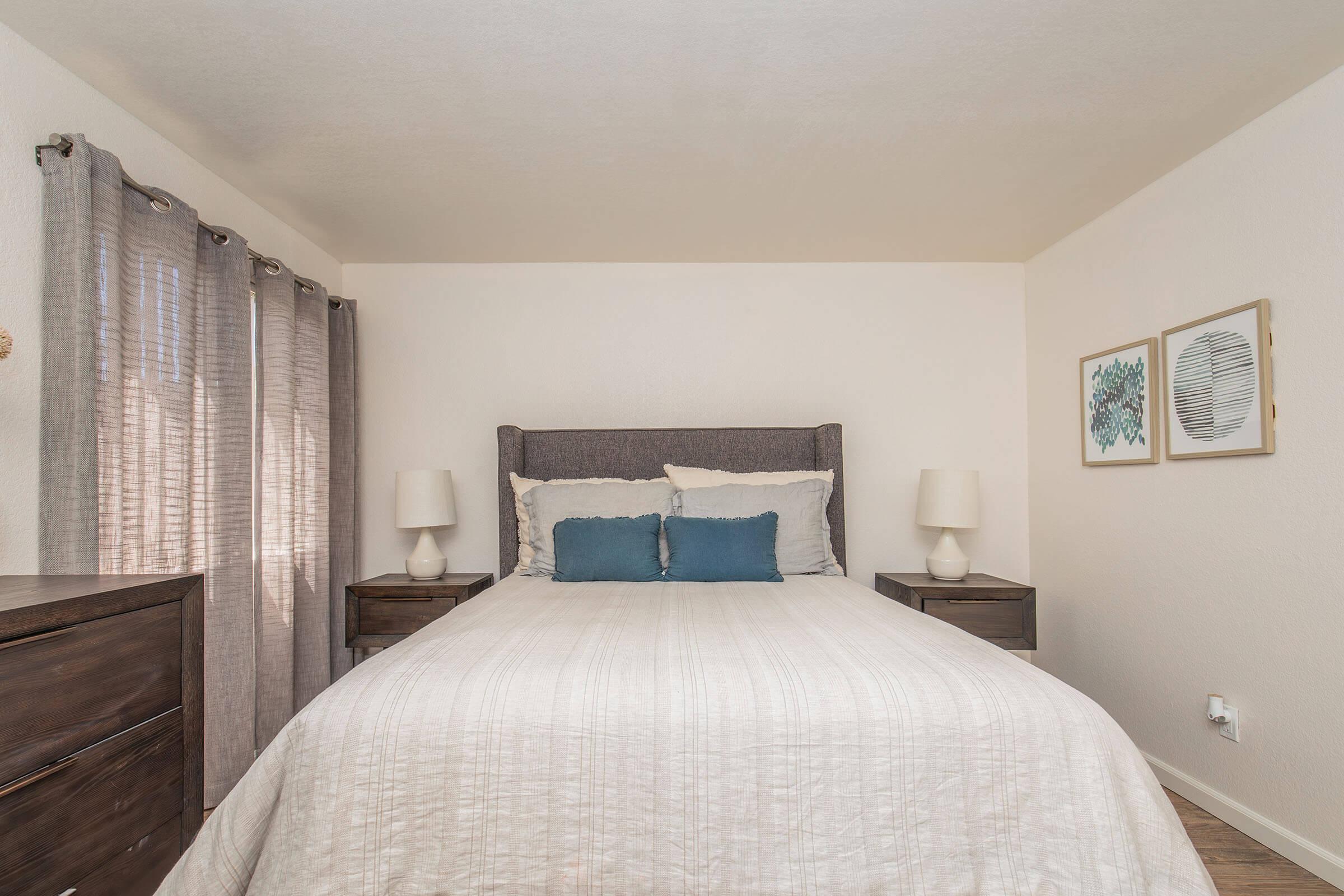 A cozy bedroom featuring a large bed with white bedding and blue accent pillows. There are two bedside tables with lamps on either side, and light gray curtains covering a window. On the wall, two framed artworks add a decorative touch to the serene space.