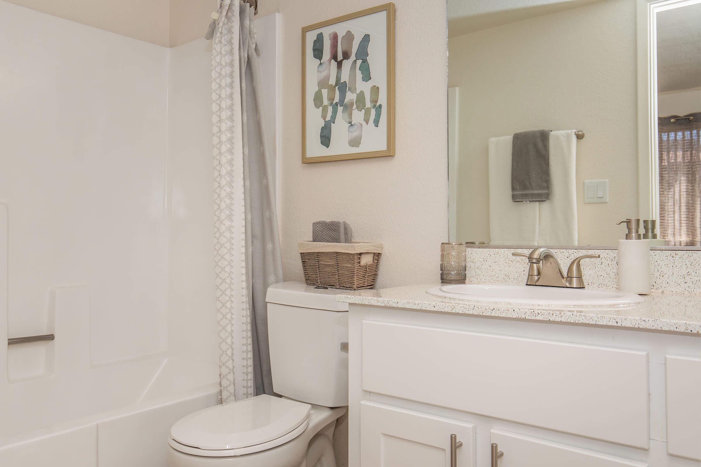 A modern bathroom featuring a bathtub with a shower curtain, a white toilet, and a vanity with a sink. The vanity has a speckled countertop, a decorative basket, and a framed artwork on the wall. Soft lighting illuminates the space, enhancing the neutral color scheme.