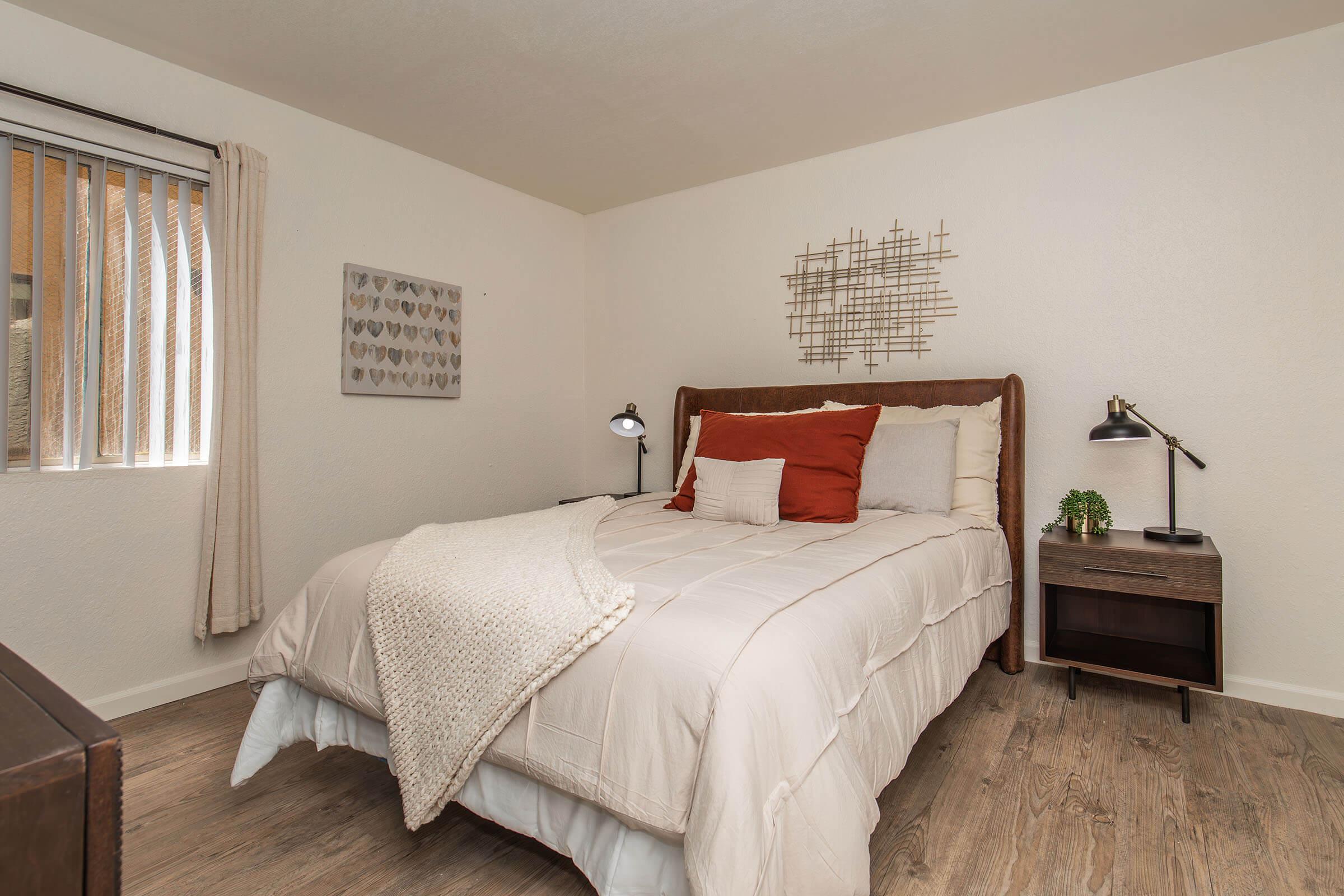 A cozy bedroom featuring a large bed with a beige comforter and decorative pillows, a wooden nightstand with a lamp, and a small potted plant. The room has light-colored walls, wooden flooring, and a decorative wall piece above the bed. Natural light comes through a window with vertical blinds.