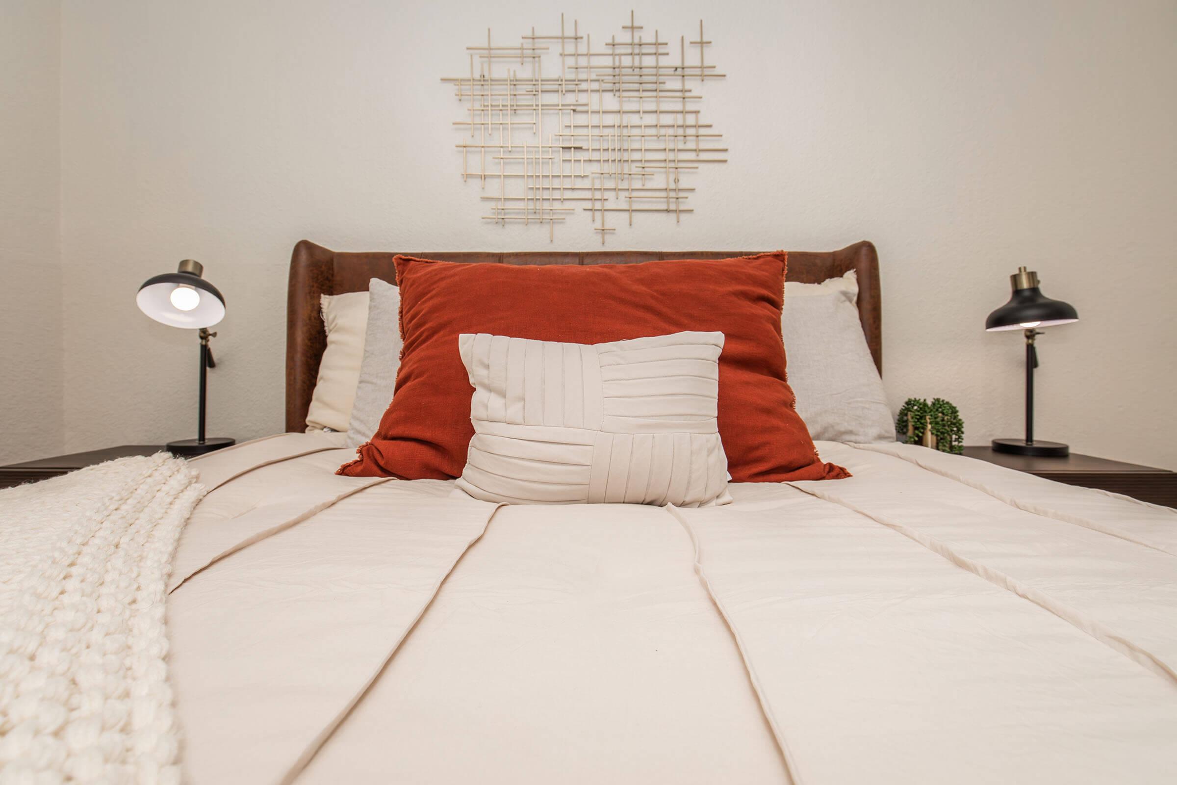 A neatly made bed with a brown headboard, featuring multiple pillows in shades of red and cream. Two modern bedside lamps sit on either side, and there is a textured white throw blanket draped over the bed. A decorative wall piece made of geometric shapes is displayed above the headboard.
