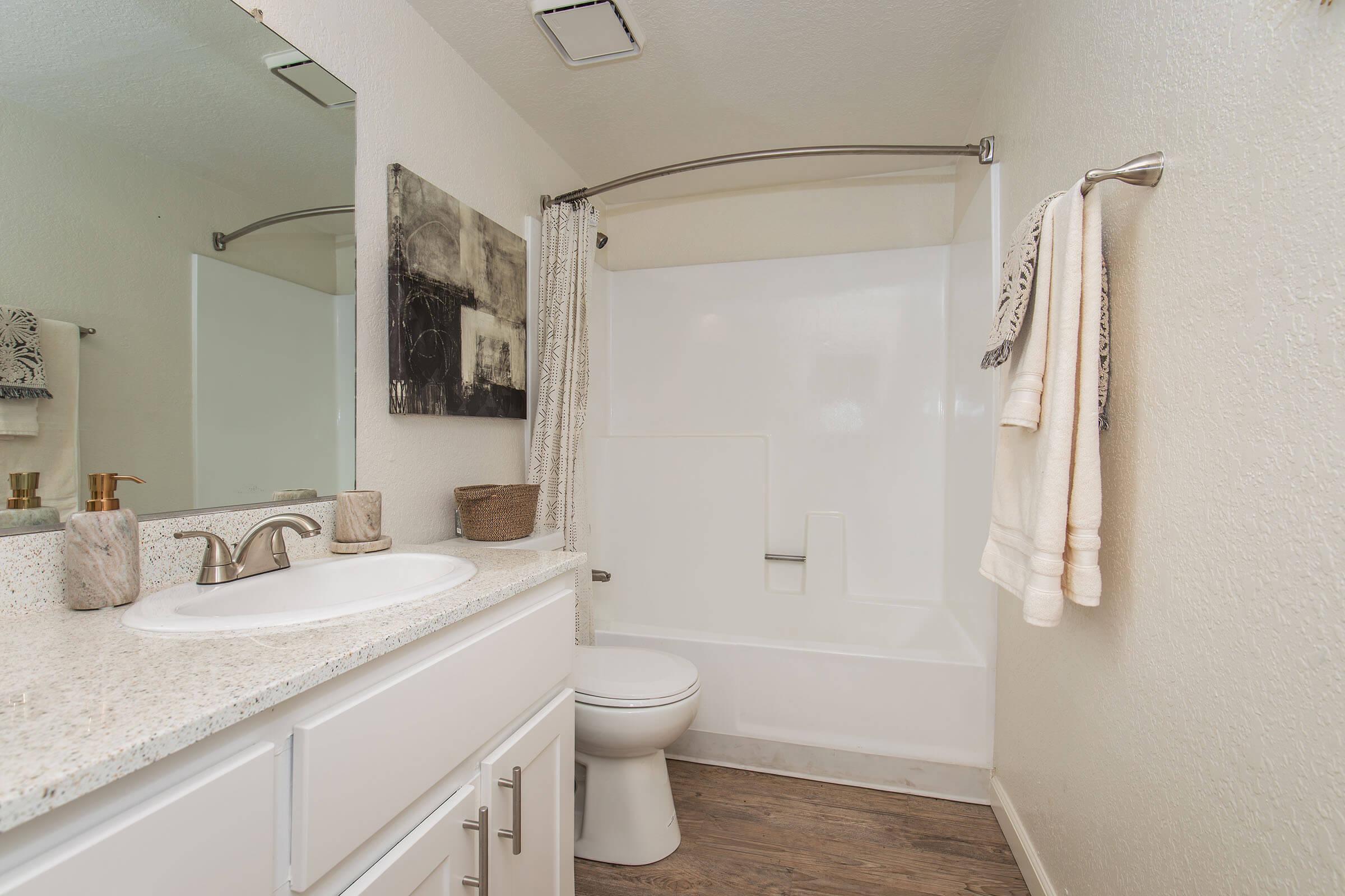 A clean, modern bathroom featuring a white tub and shower combo. The vanity has a rectangular sink, a large mirror above, and is adorned with decorative items. Soft beige towels hang on a rack, and there are neutral-toned walls and flooring, creating a bright and inviting atmosphere.