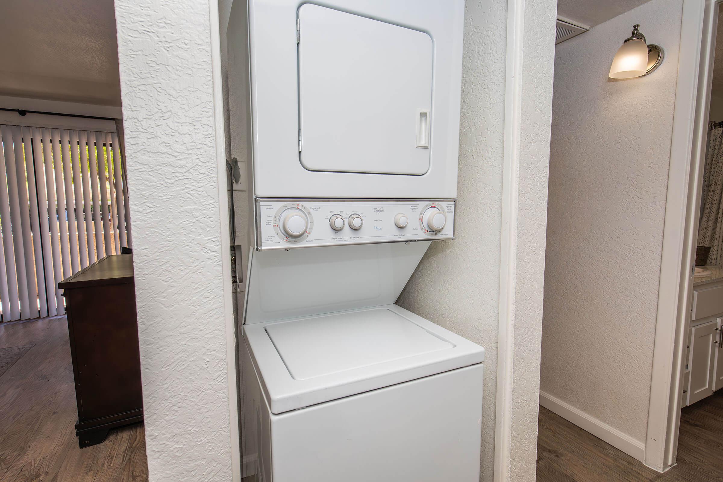 A stacked washer and dryer unit is located between two walls in a brightly lit space. The appliances are white with control dials and buttons clearly visible. The area has light-colored walls and wood-like flooring, creating a clean and modern appearance. Bright daylight filters through nearby blinds.