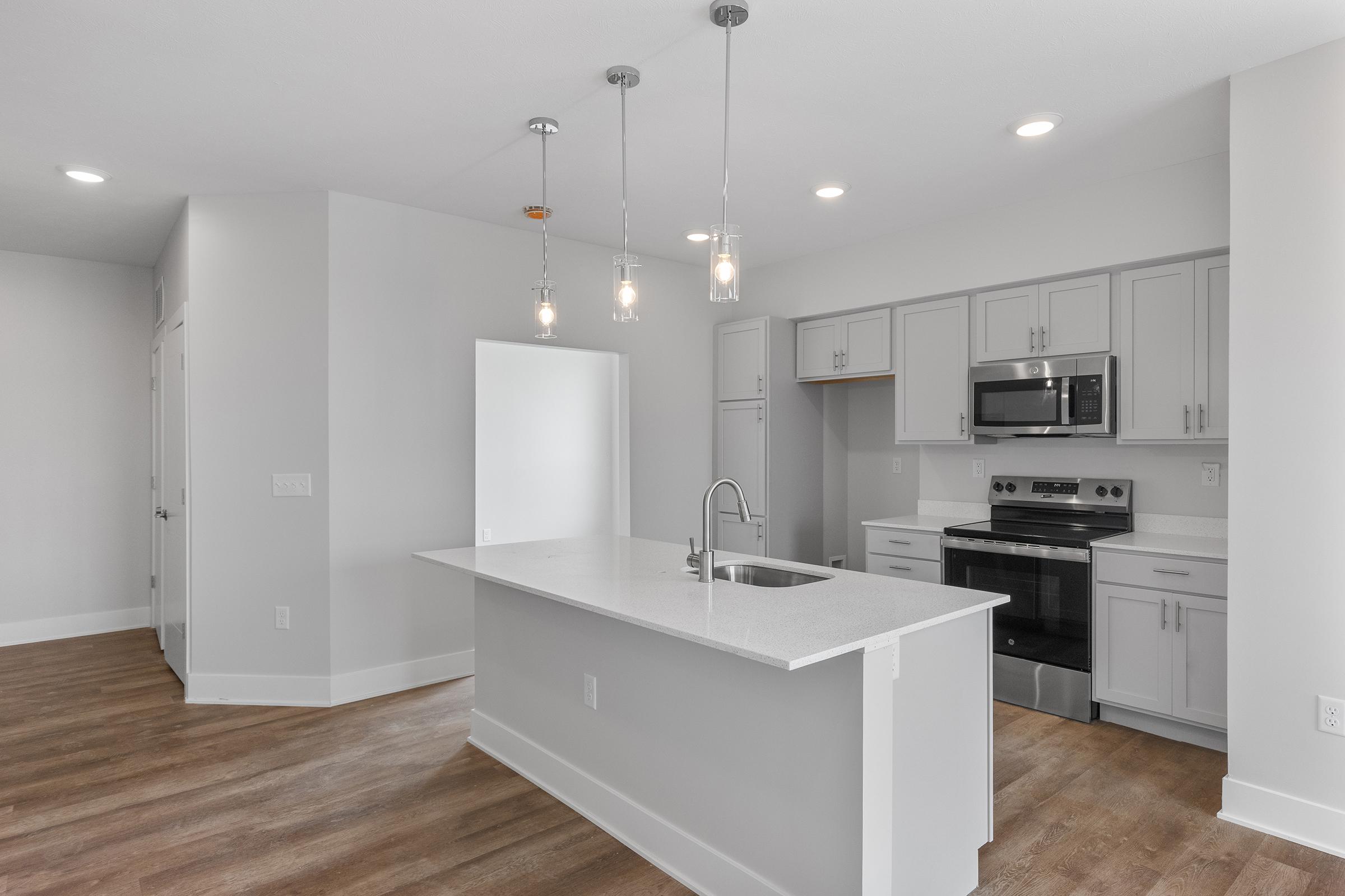 A modern kitchen featuring a central island with a sink, stainless steel appliances, and pendant lighting. The cabinetry is light gray, complemented by a white countertop. The floor has a warm wooden finish, and the space is well-lit with natural light coming from a nearby doorway.