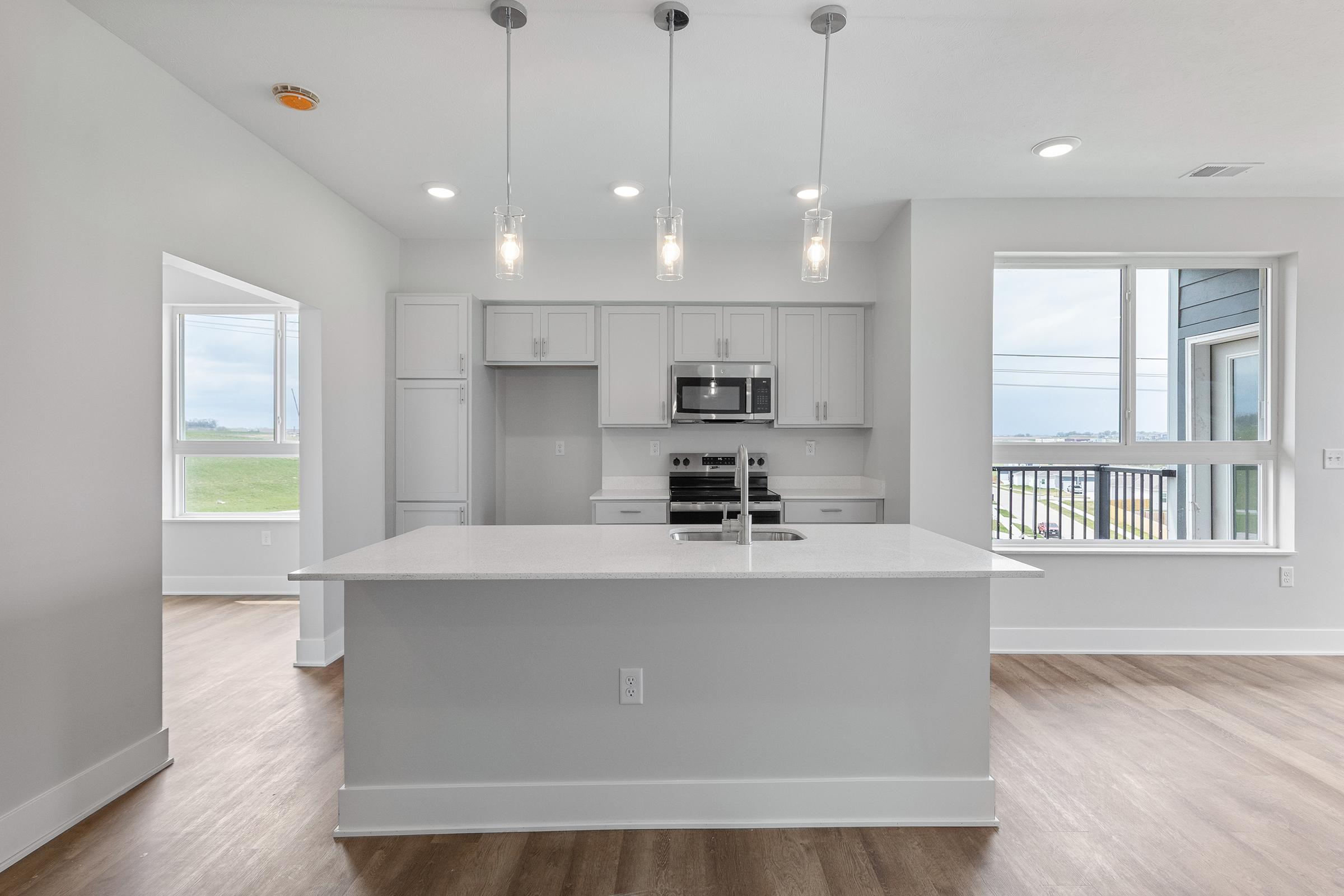 Modern kitchen interior featuring an island with a sink, light gray cabinetry, stainless steel appliances including a microwave and oven, and pendant lighting. Large windows allow natural light and showcase a view of the outdoors. The floor is a light wood finish, creating a bright and airy atmosphere.