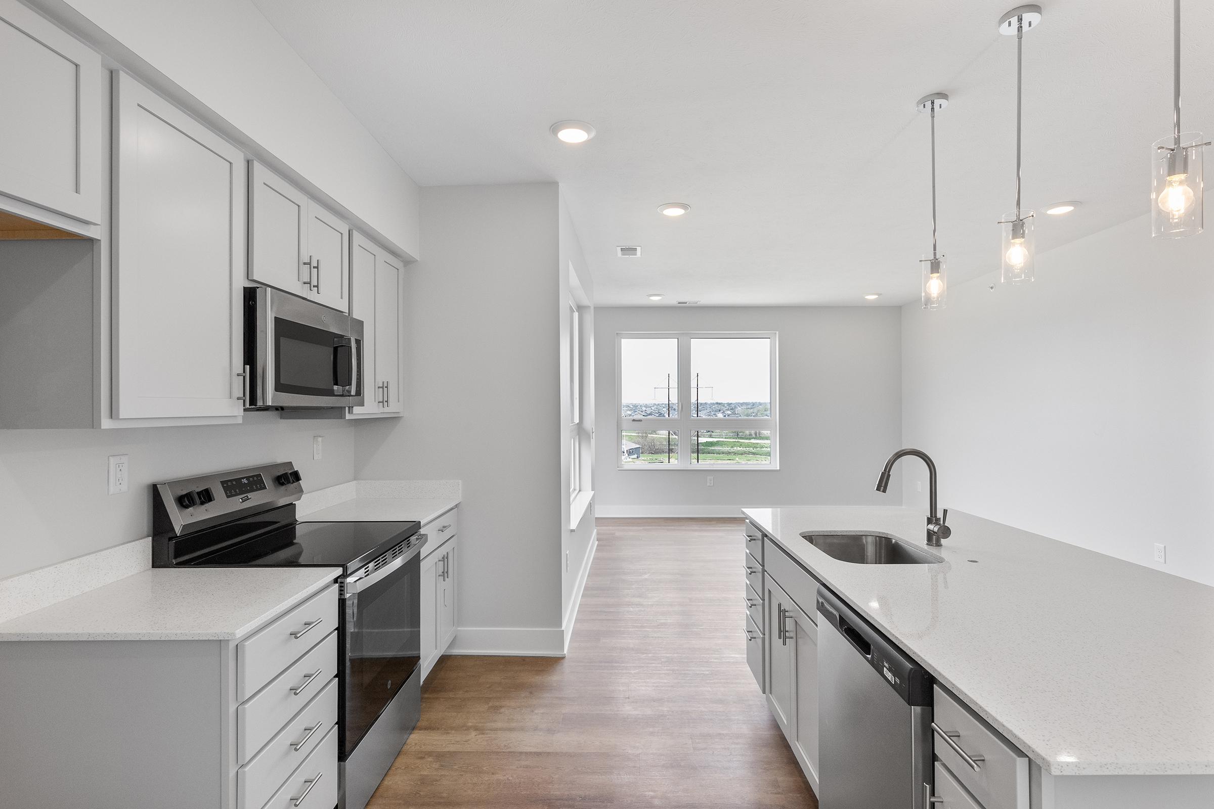 Modern kitchen with gray cabinets, stainless steel appliances including a microwave and oven, a large island with a sink, and pendant lighting. The space features hardwood flooring and large windows that allow natural light, providing a view of the outdoors.