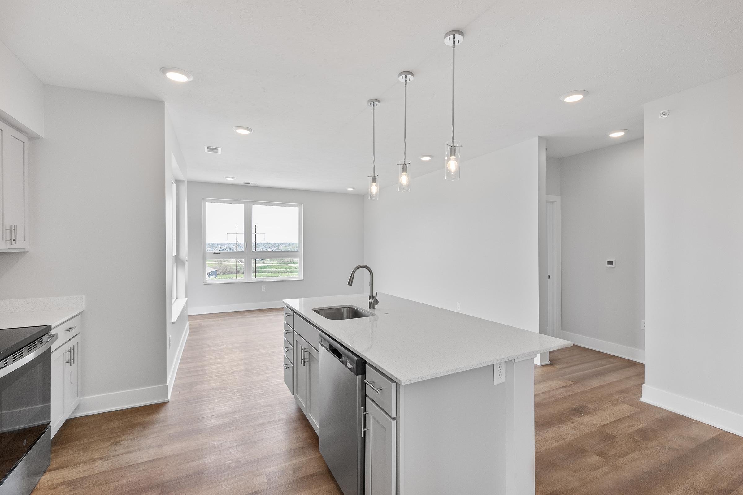 Modern kitchen and living area featuring a central island with a sink, stainless steel dishwasher, and pendant lighting. Large windows provide natural light and a view of the outdoors, with light-colored walls and wood flooring creating a fresh and airy atmosphere.