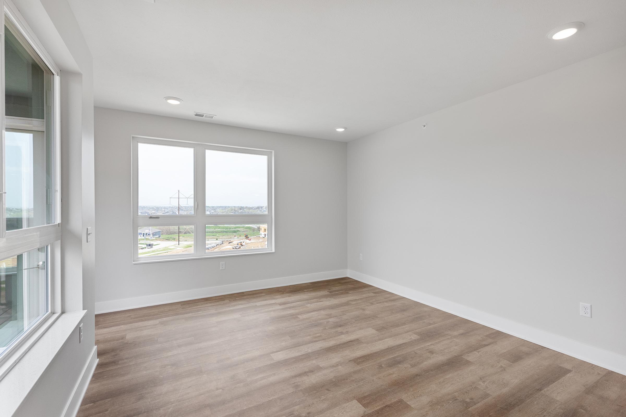 Spacious interior of a modern room featuring large windows that provide natural light, light-colored walls, and new wooden flooring. The room is empty, giving a sense of openness and potential for personalization.