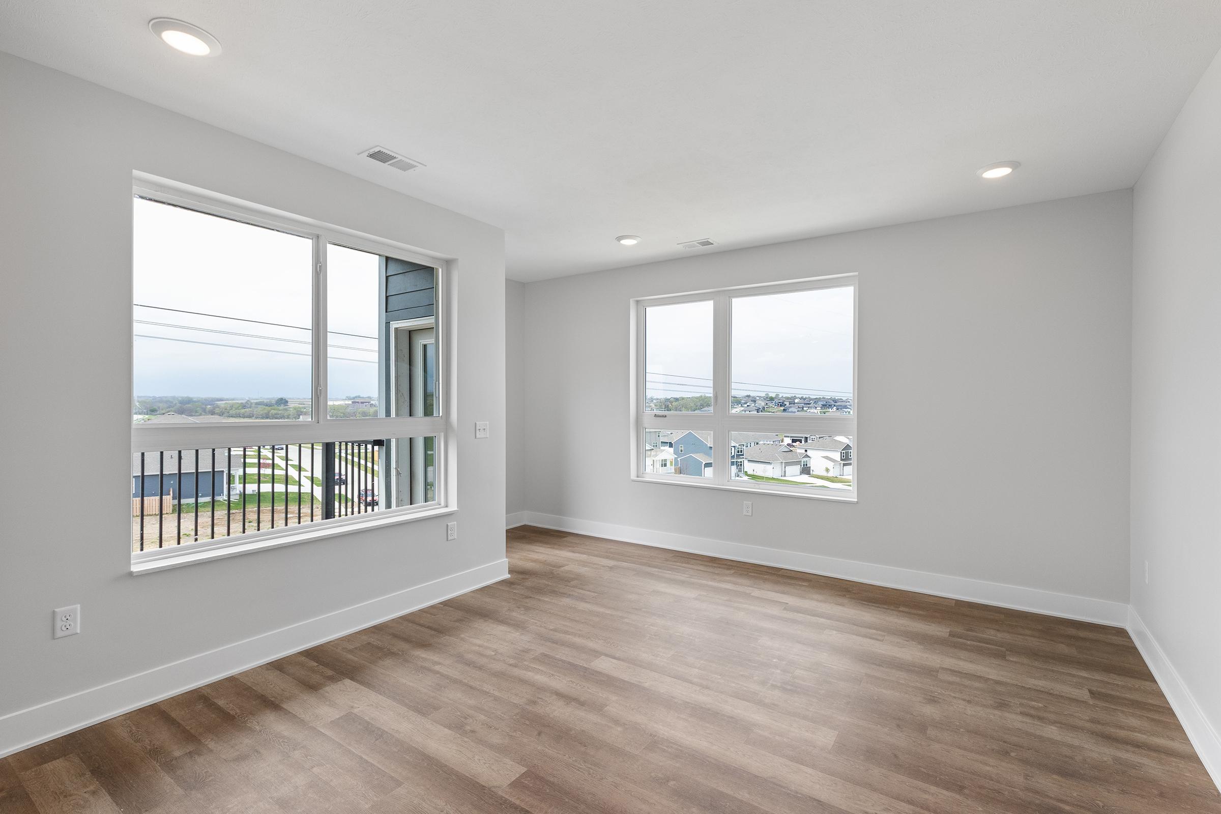 A bright and empty living room featuring large windows that provide a view of the outdoors. The walls are painted a light color, and the floor is covered with laminate wood. Recessed lighting is installed in the ceiling, enhancing the open and airy atmosphere of the space.