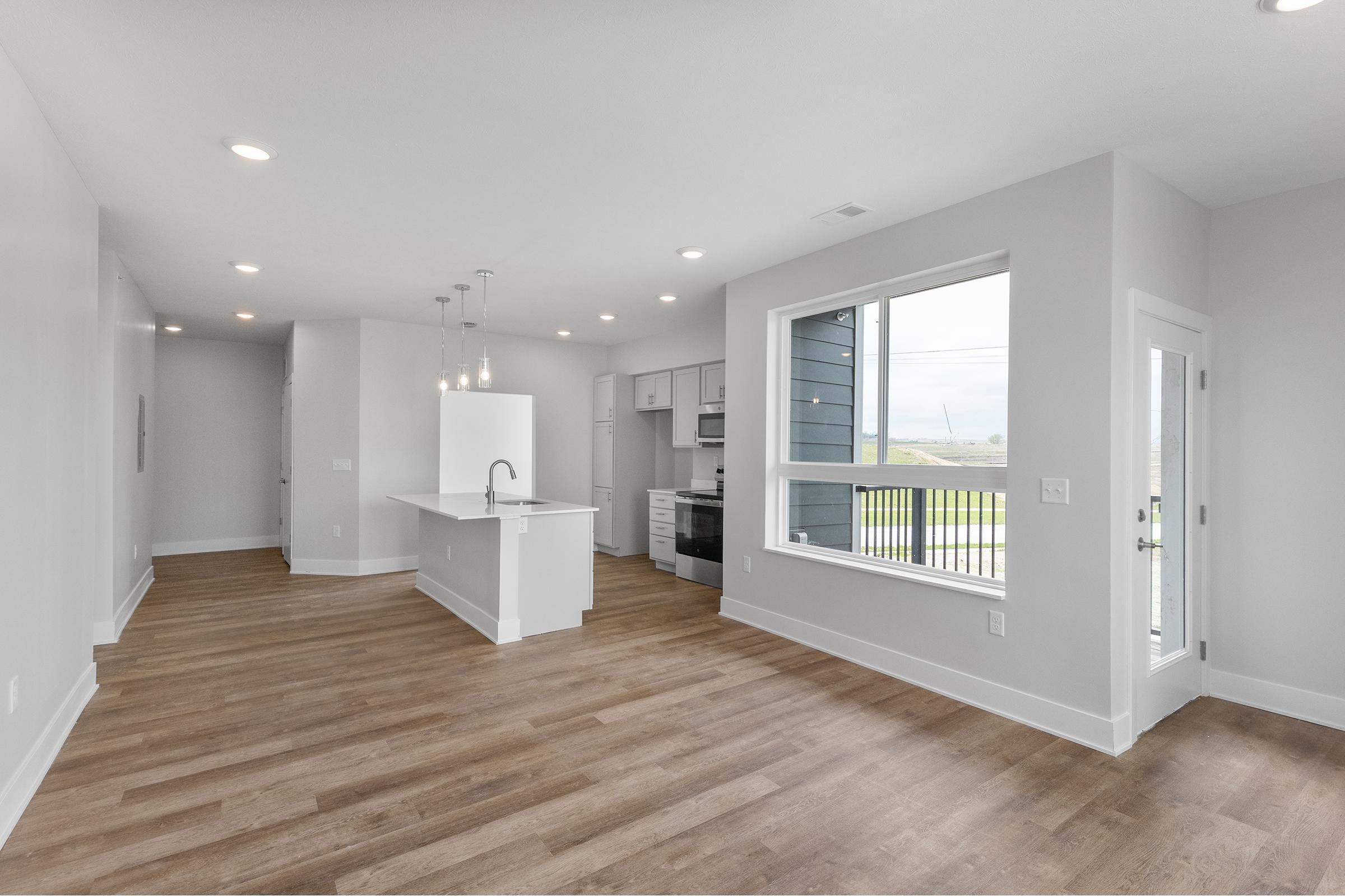 Modern open concept living space featuring a kitchen with a white island and sleek appliances, light gray walls, and large windows allowing natural light. The flooring is wood-like, and the room includes recessed lighting. A balcony can be seen through the window, enhancing the airy feel of the space.