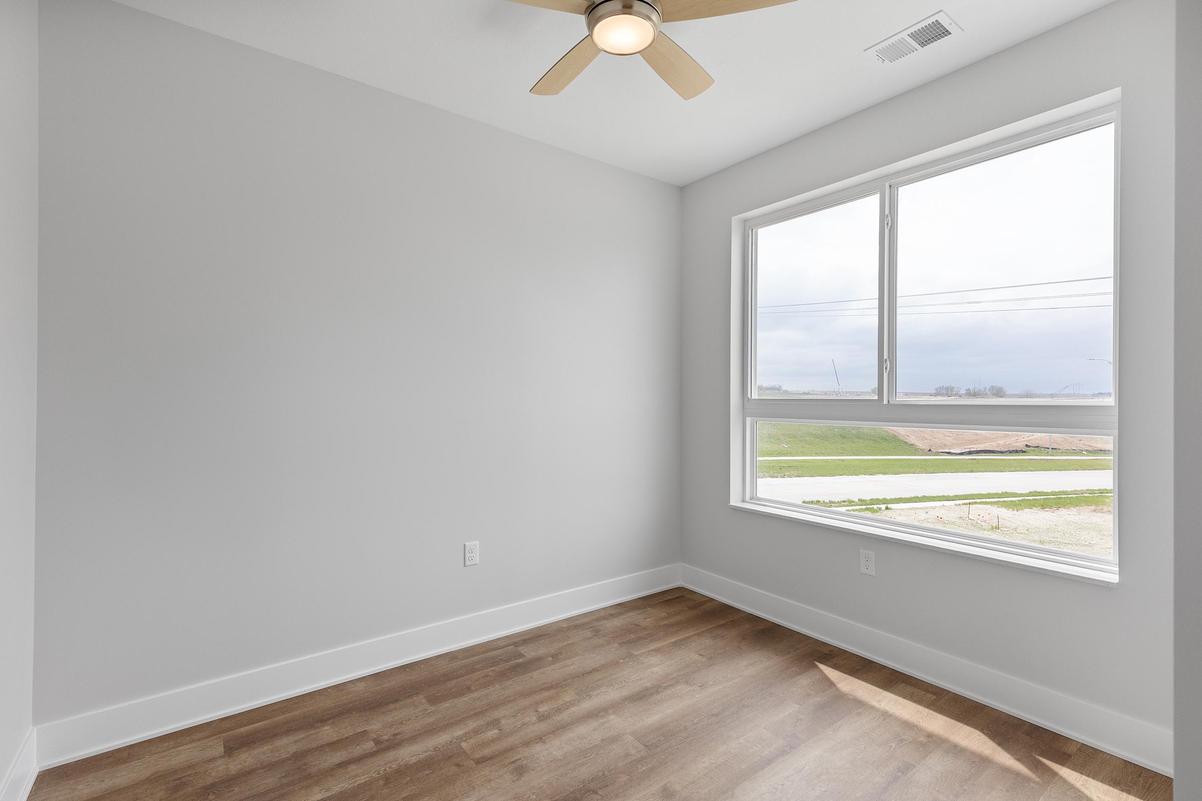 A bright, empty room with light gray walls and a ceiling fan. There is a large window that offers a view of a green landscape outside. The floor features light wood laminate. No furniture is present, giving the space a clean and minimalist appearance.