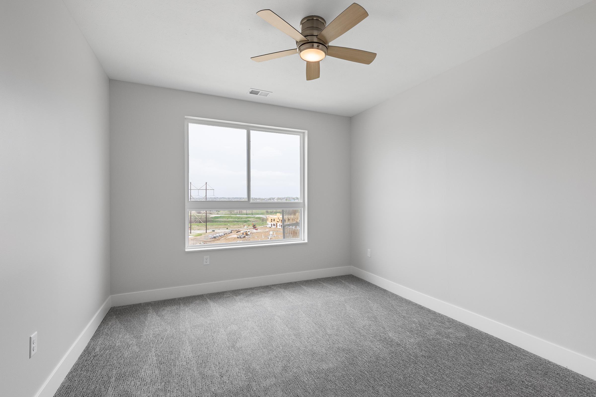 A bright, empty room featuring light gray walls and a large window with a view. The floor is covered in plush gray carpet, and a modern ceiling fan is mounted above. The space is well-lit by natural light coming through the window, creating an open and airy atmosphere.