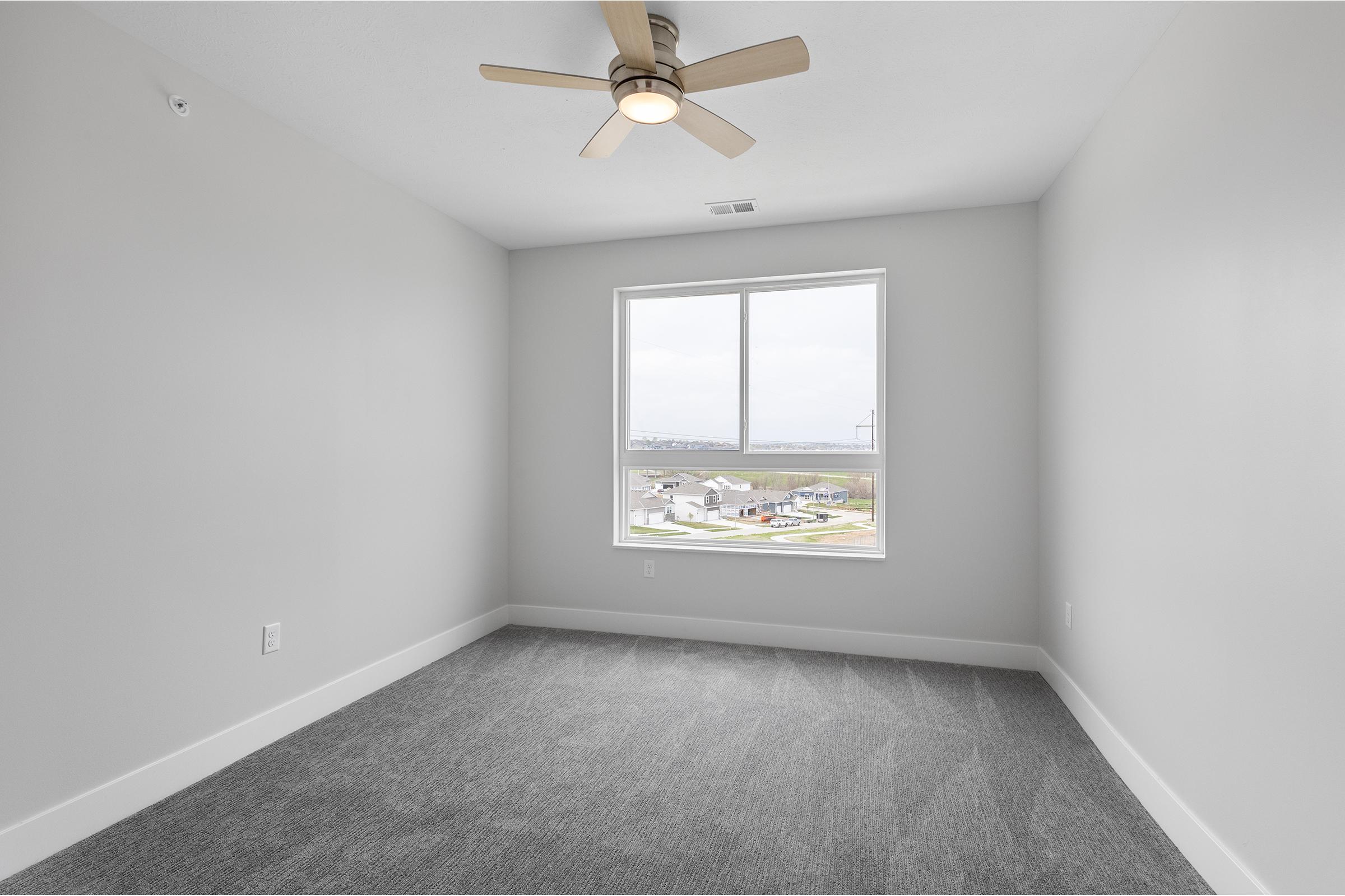 Empty room with gray walls and carpet, featuring a single window with a view. A ceiling fan is installed, and the space appears bright and airy. The room is unfurnished, providing a blank canvas for decoration or use.