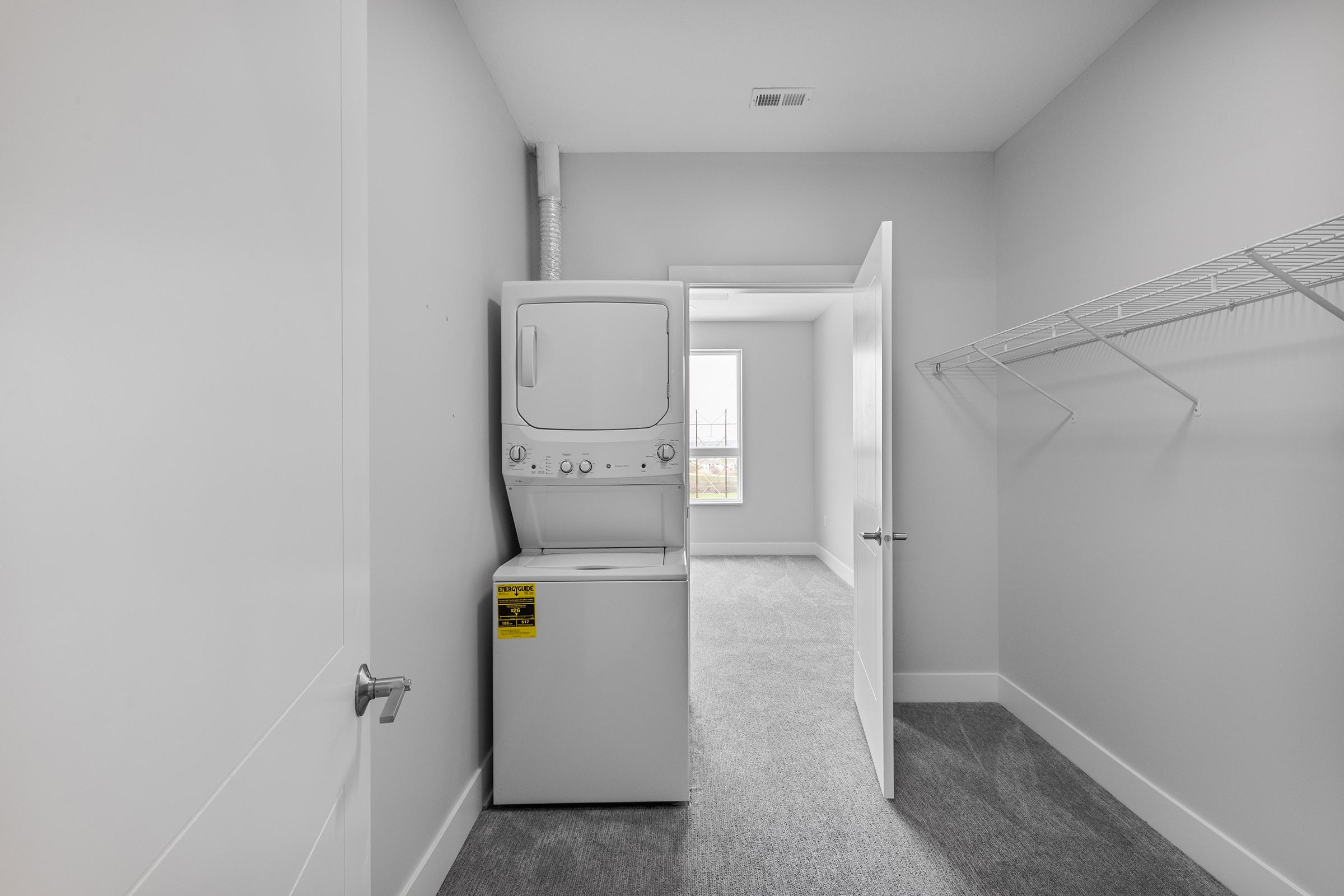 A laundry room featuring a stacked washer and dryer set against a light gray wall. A door is partially open, revealing a bright room with a window and carpeted floor. An empty wire shelf hangs on the wall to the right.