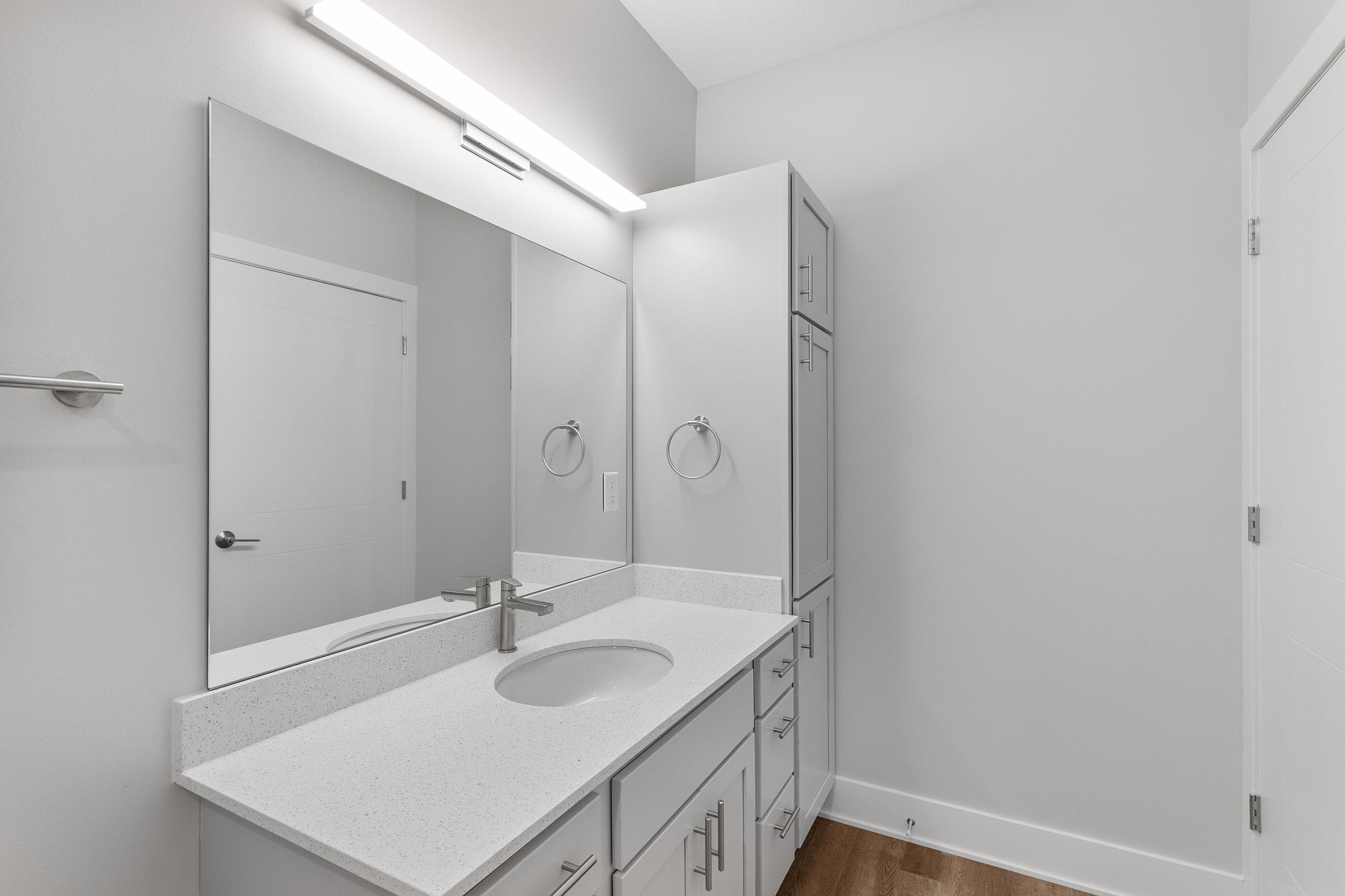 Modern bathroom interior featuring a large mirror above a white countertop with a single sink. The cabinetry is light-colored and sleek, complementing the neutral wall tones. There is a towel ring and a door visible, suggesting a clean and minimalist design. Good lighting from a fixture above the mirror.
