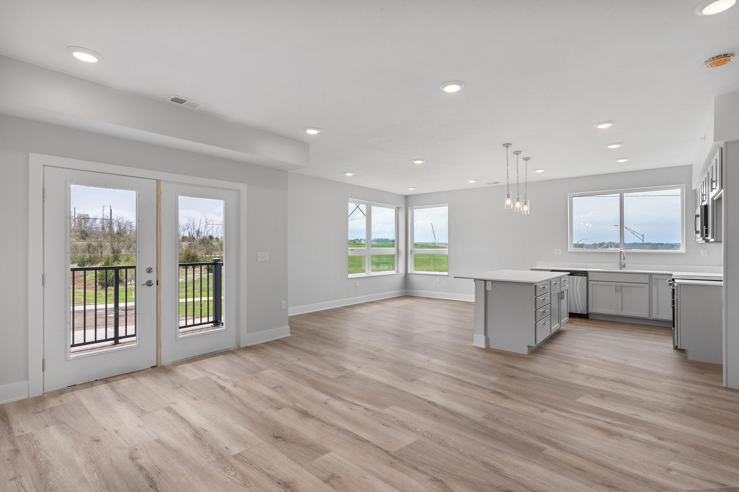 Bright and spacious interior of a modern living area with hardwood floors, large windows providing natural light, and a stylish kitchen featuring an island. Doors lead to a balcony with a view of green fields. The decor is minimalistic and contemporary, creating an inviting atmosphere.
