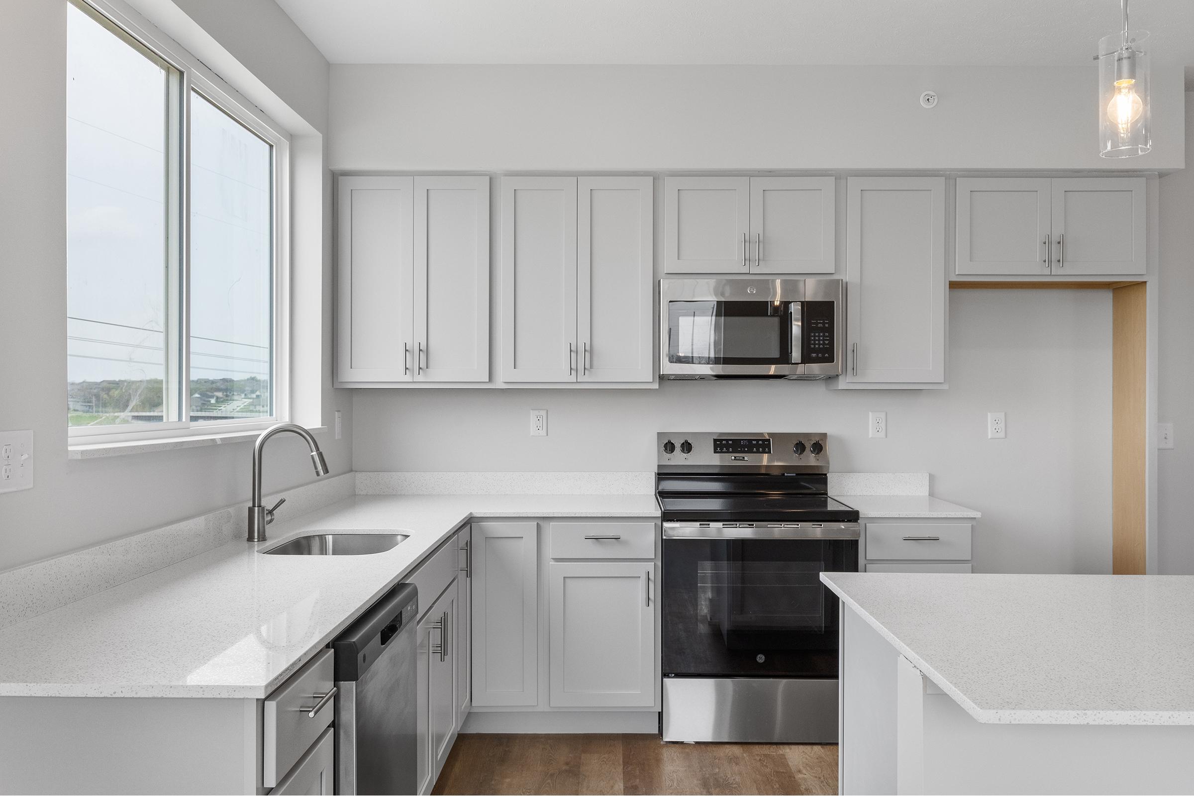 Modern kitchen featuring light gray cabinetry, stainless steel appliances including a microwave and oven, and quartz countertops. Large window providing natural light, with an open layout that connects to a nearby room. Wood flooring adds warmth to the space.