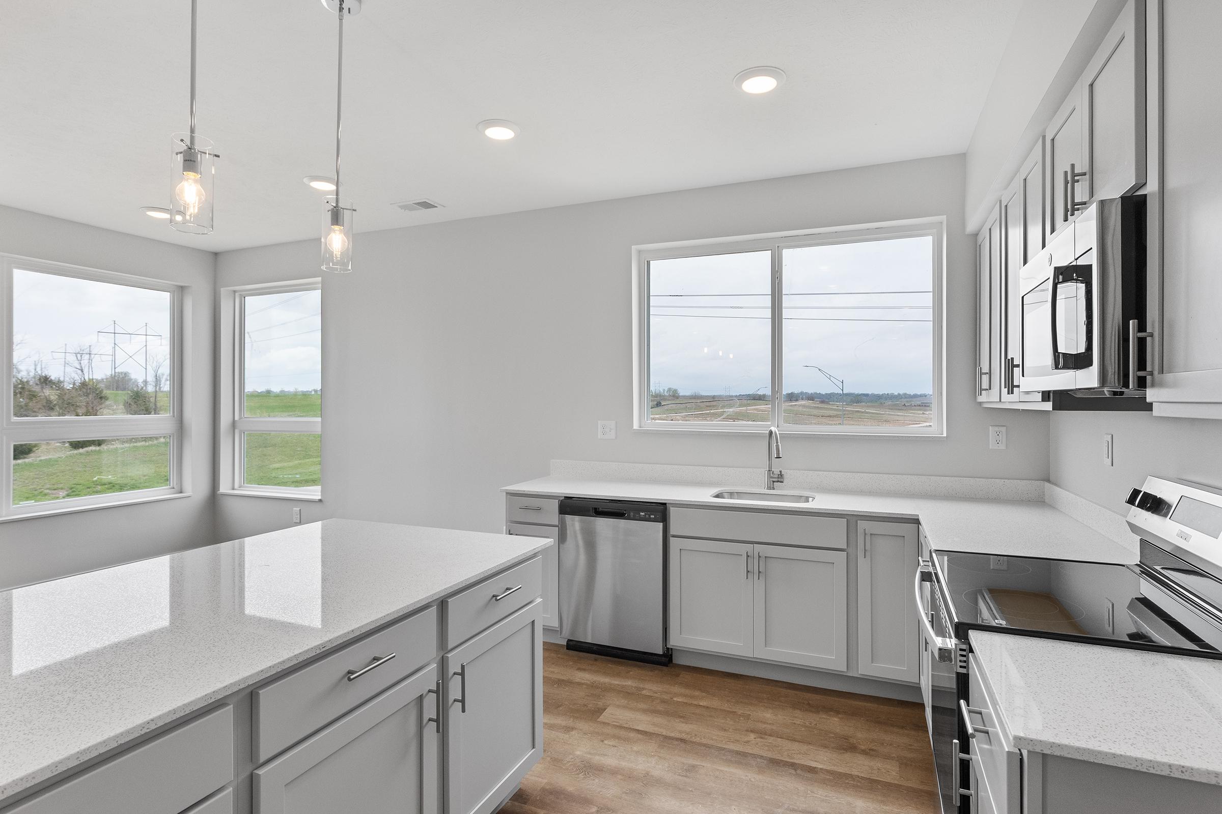 Modern kitchen featuring light grey cabinetry, stainless steel appliances, and a large island with a white quartz countertop. Large windows allow natural light and provide views of the surrounding area. The flooring is a light wood finish, enhancing the bright and airy atmosphere.