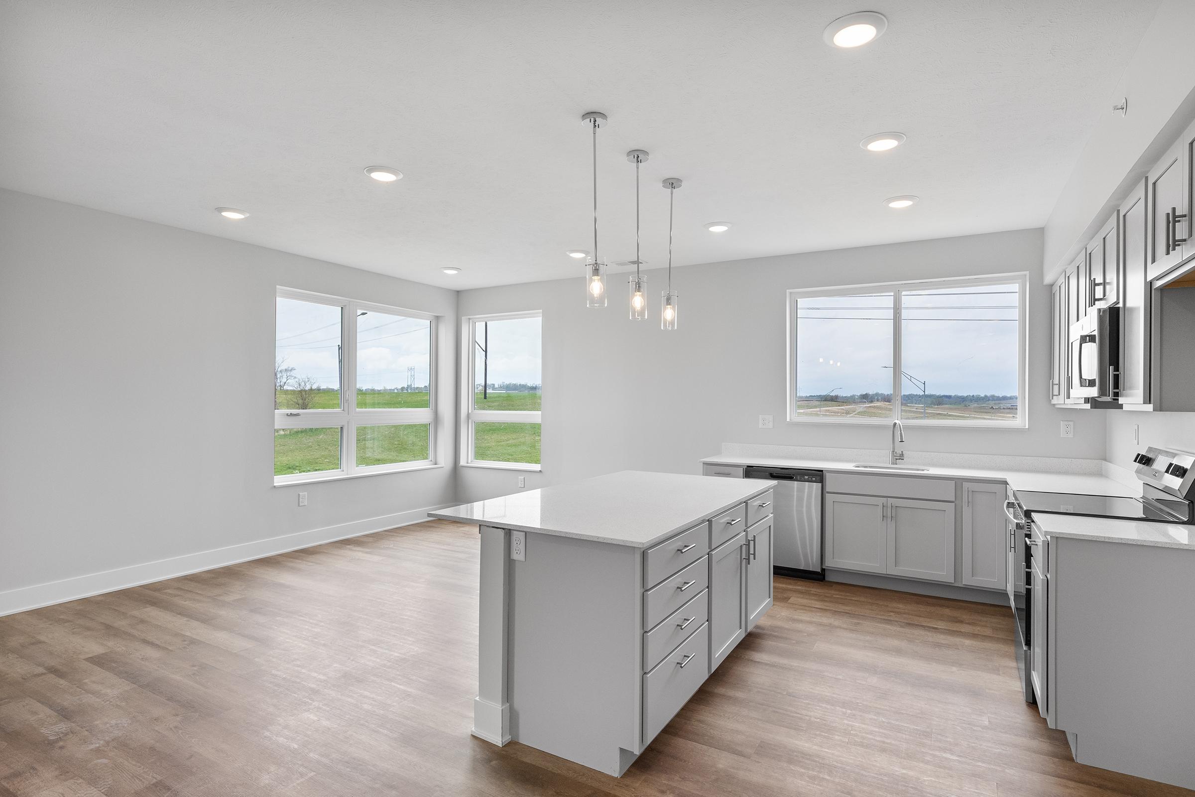 Modern kitchen with a large island and white cabinetry. Brightly lit with pendant lights, large windows offer natural light and a view of the greenery outside. The flooring is wood, complementing the contemporary design. Stainless steel appliances and sleek countertops enhance the space's functionality and style.
