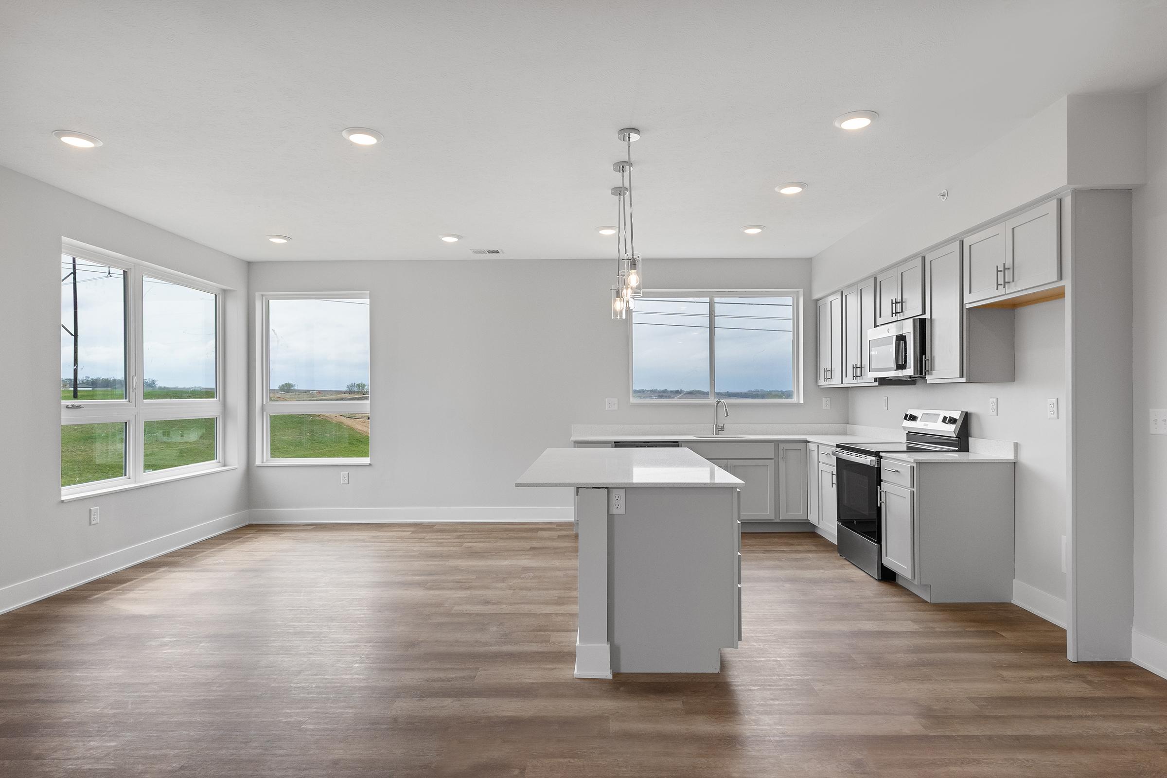 Modern open-concept kitchen and living area featuring a central island with seating, stainless steel appliances, and ample natural light from large windows. The floor is made of light wood, creating a bright and airy atmosphere. A scenic view of the outdoors is visible through the windows.