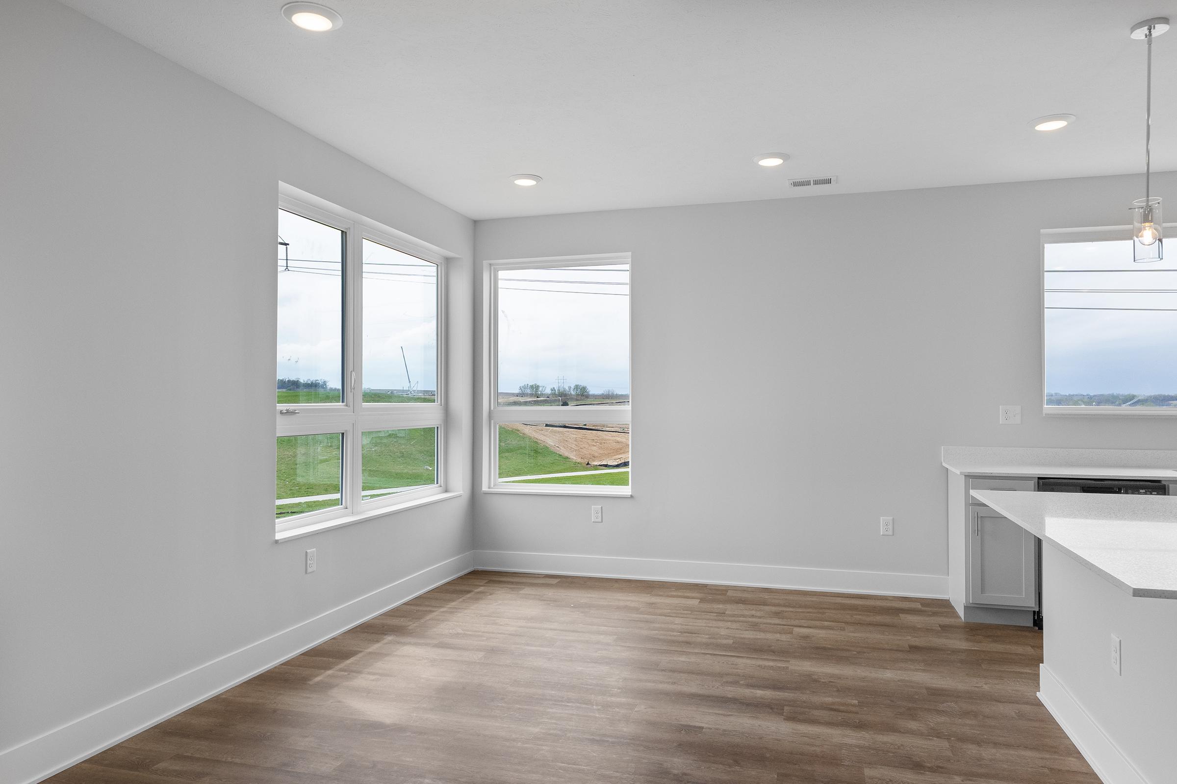 A bright, modern interior of a room featuring large windows that offer a view of the outdoors. The walls are painted white, and the flooring is light wood. There’s a kitchen area in the corner with a white countertop, providing a spacious and open feel to the living space.